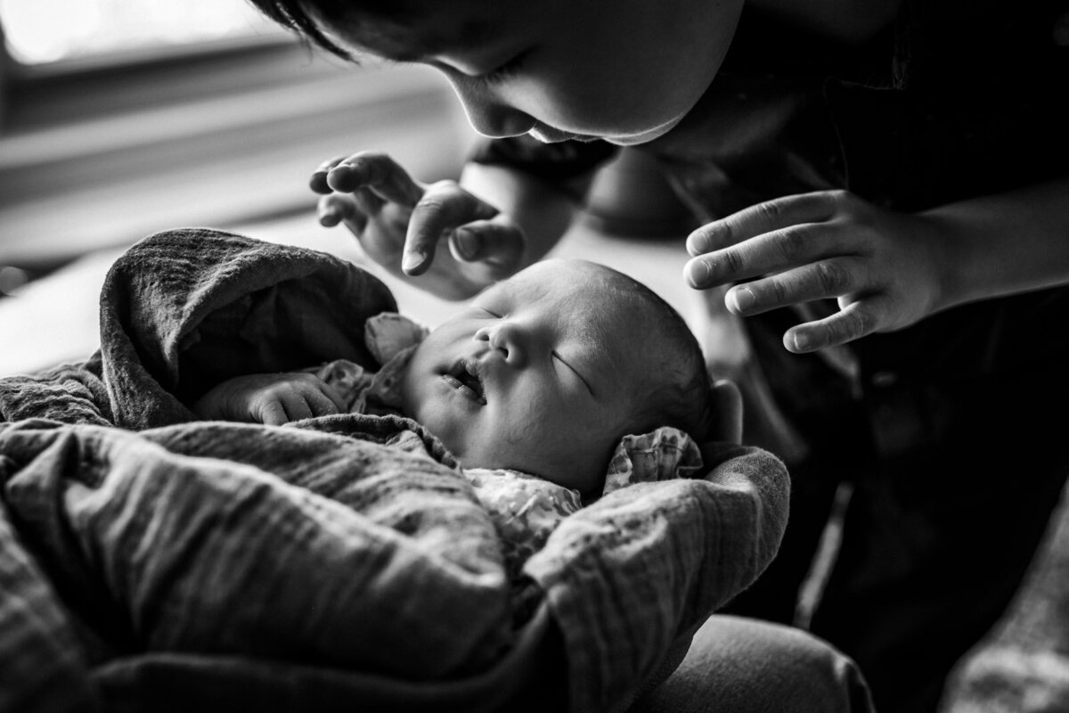 brother leaning over swaddled sleeping newborn and reaching for her head
