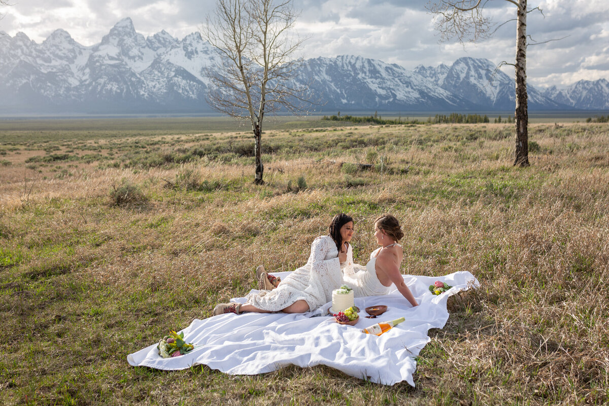 Two brides sit on a blanket in the grass sharing a picnic on their elopement day in Wyoming.