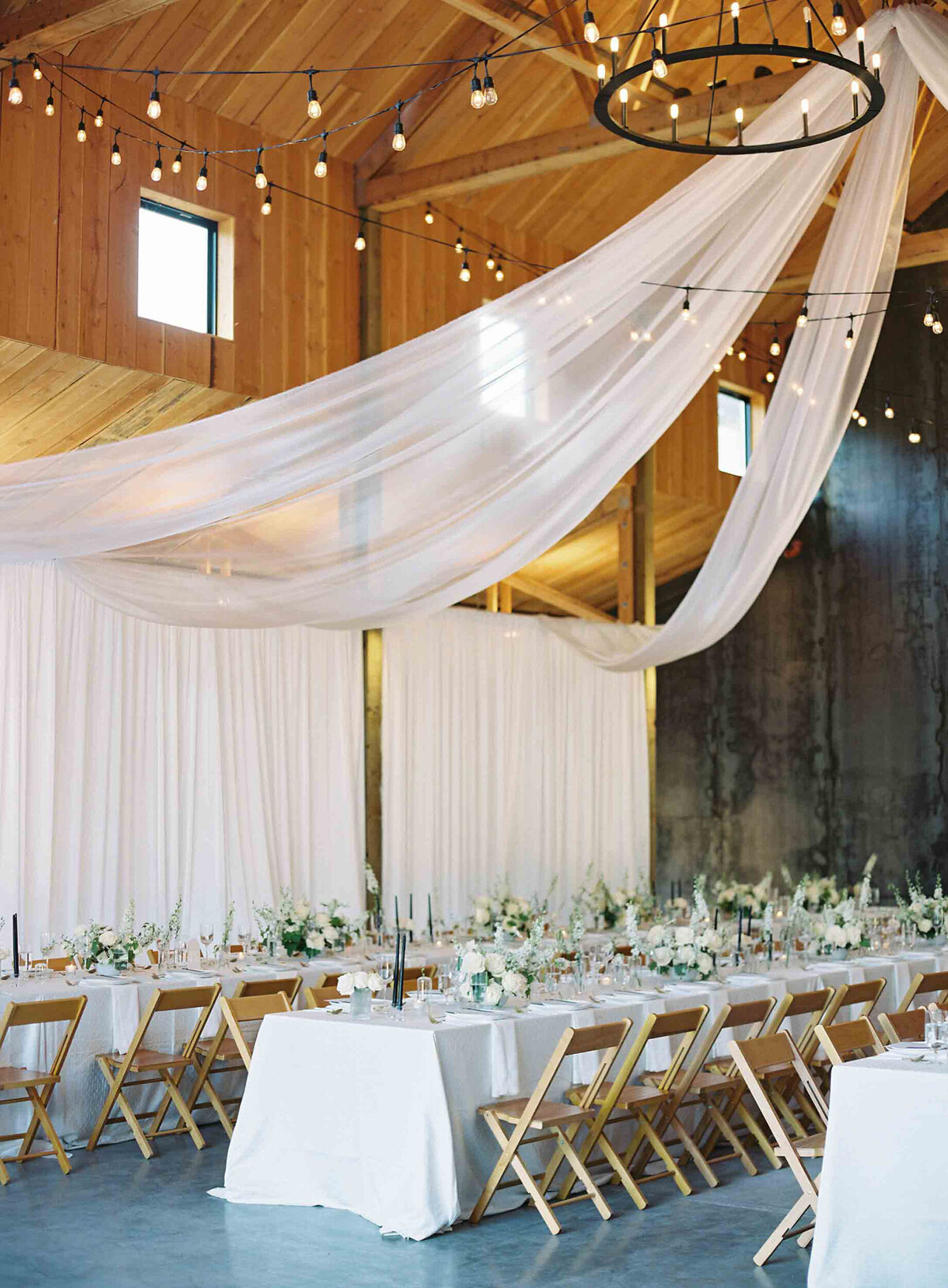 barn wedding reception at saltwater farms with white draping hanging from ceiling and wooden folding chairs and white linens with green florals