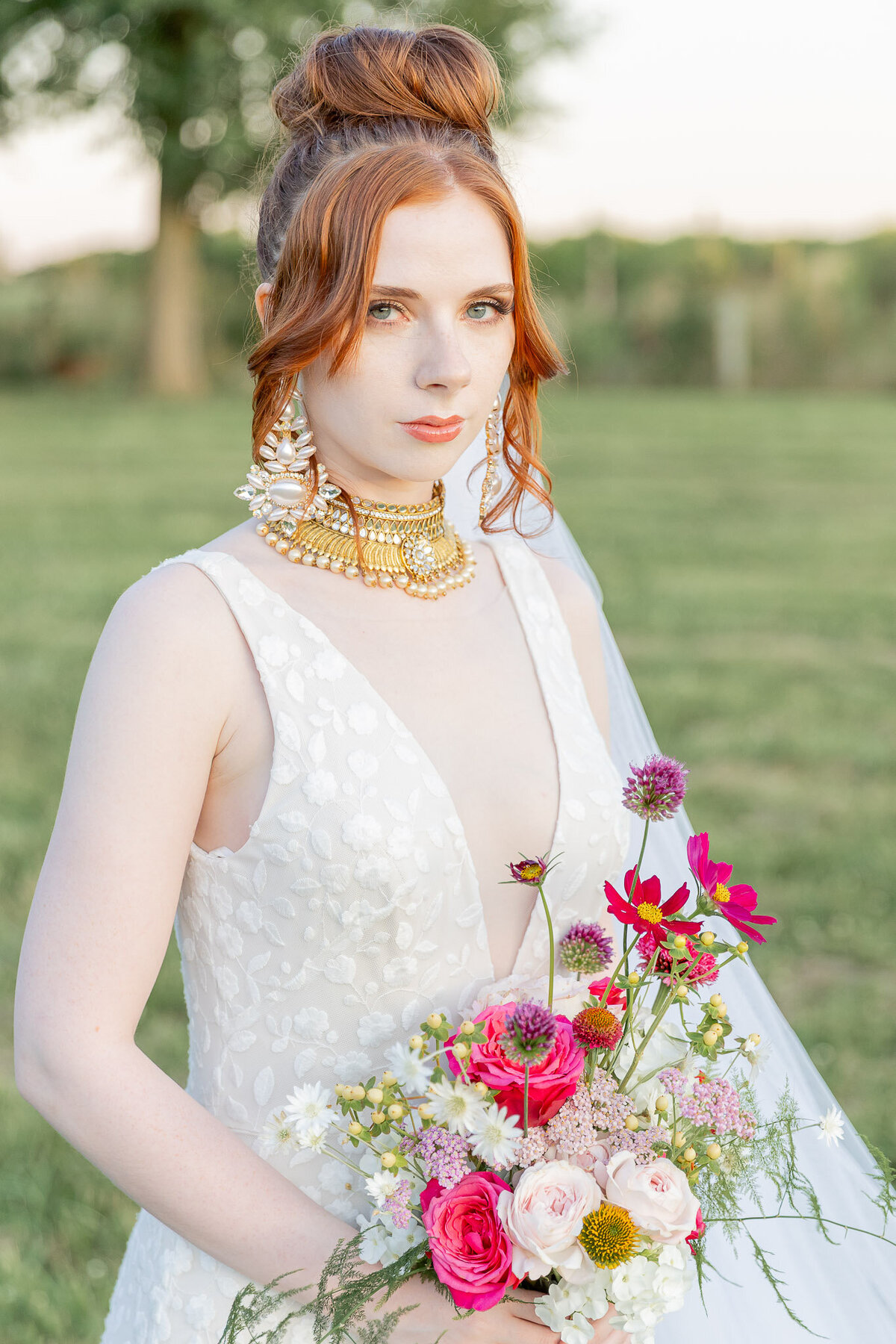 A portrait of a bride looking at the camera while wearing large, chunky jewelry along with her wedding dress. She's holding a bouquet of bright pink flowers.