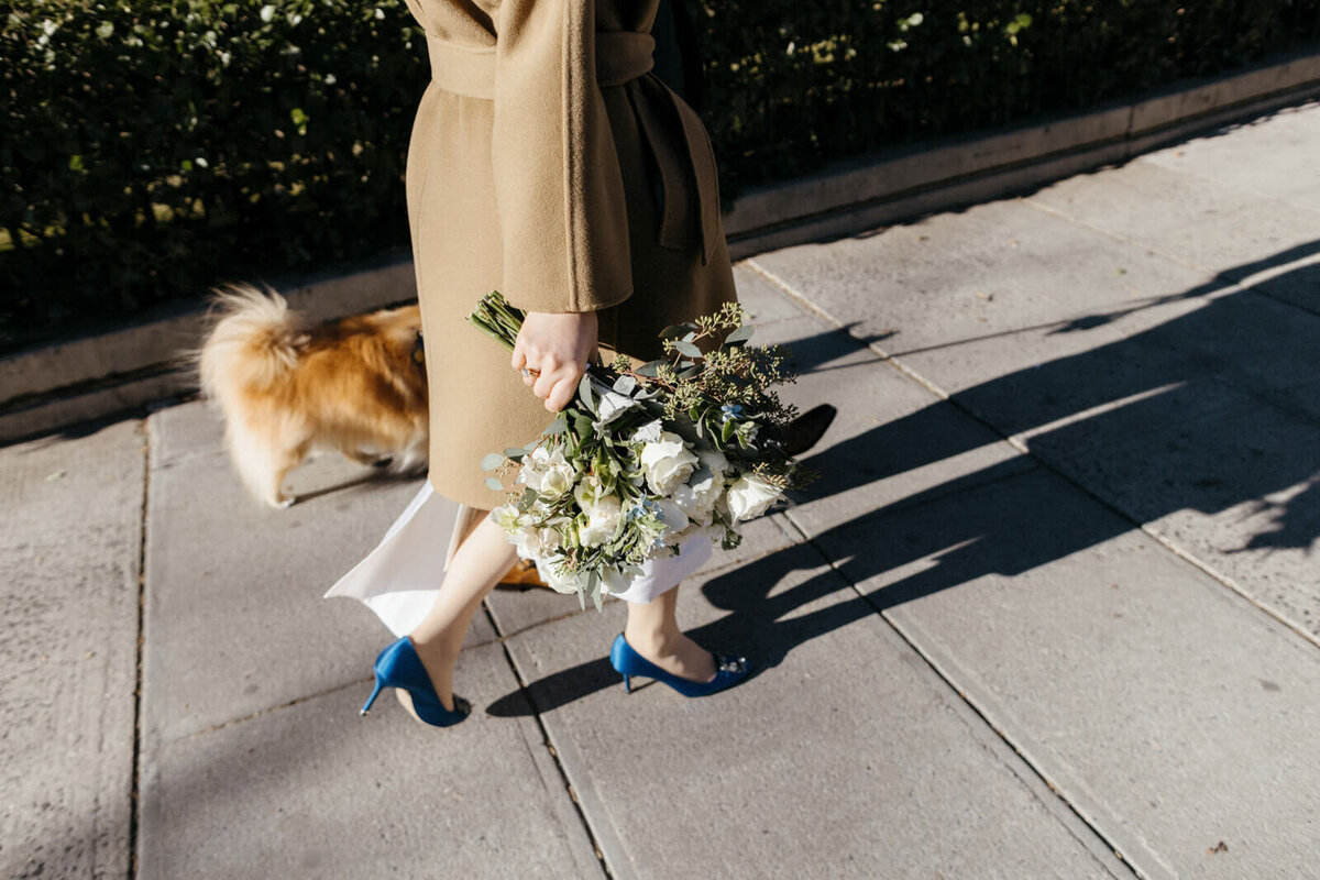 Half-body shot of the bride, who is wearing a brown coat and blue shoes, holding her flower bouquet while walking with her dog.