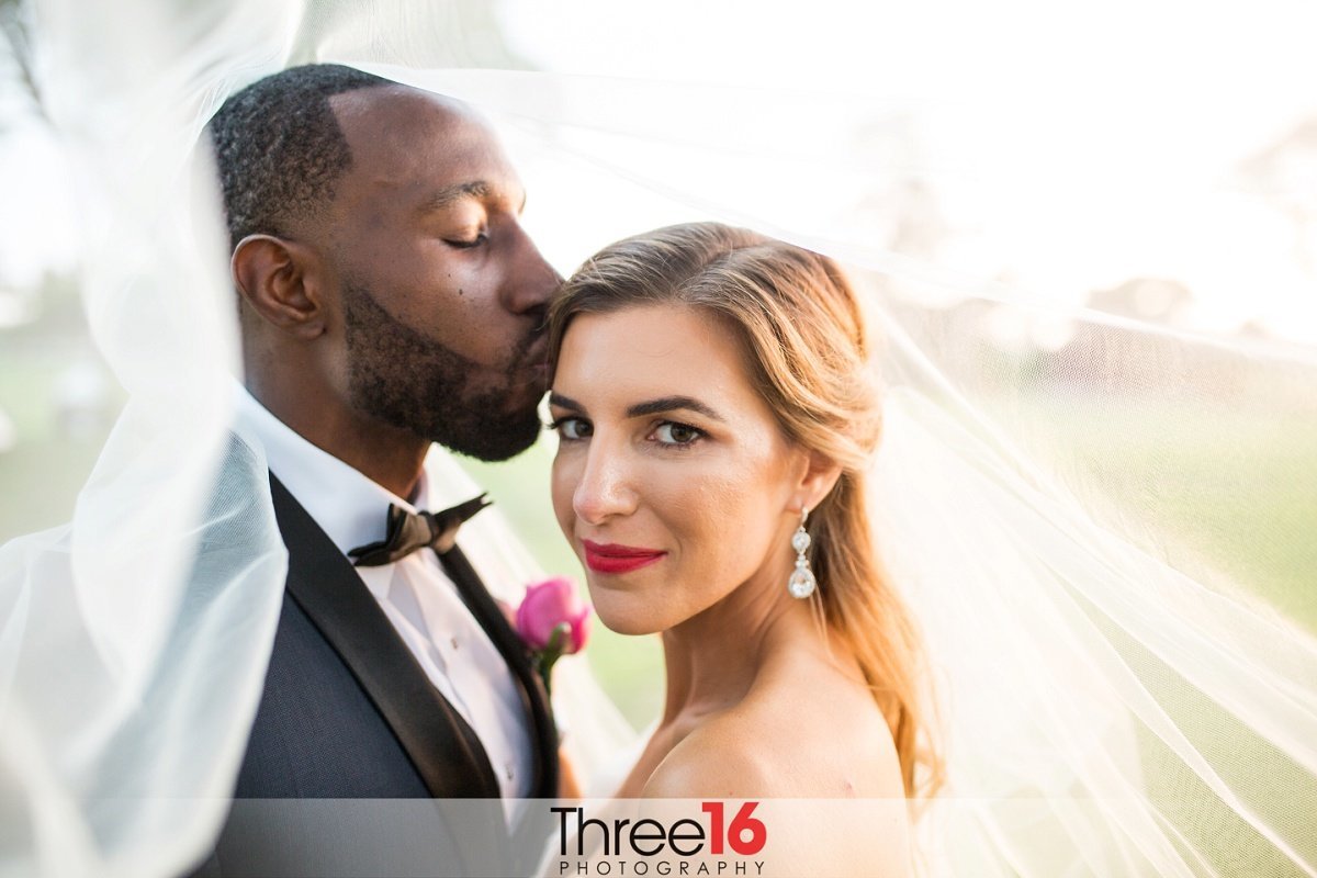Groom kisses his Bride on the side of her head as she looks at the camera