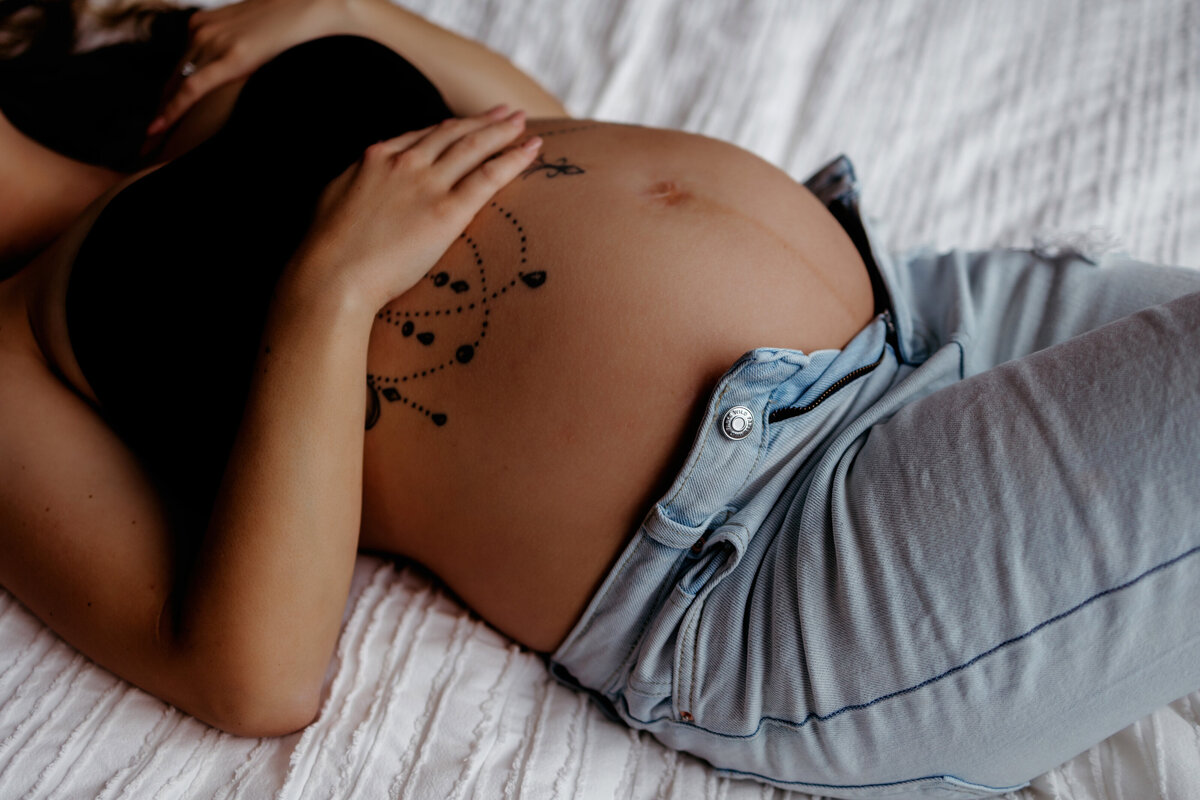 pregnant mom lays on bed in black bra and jeans with tatoos on pregnant belly