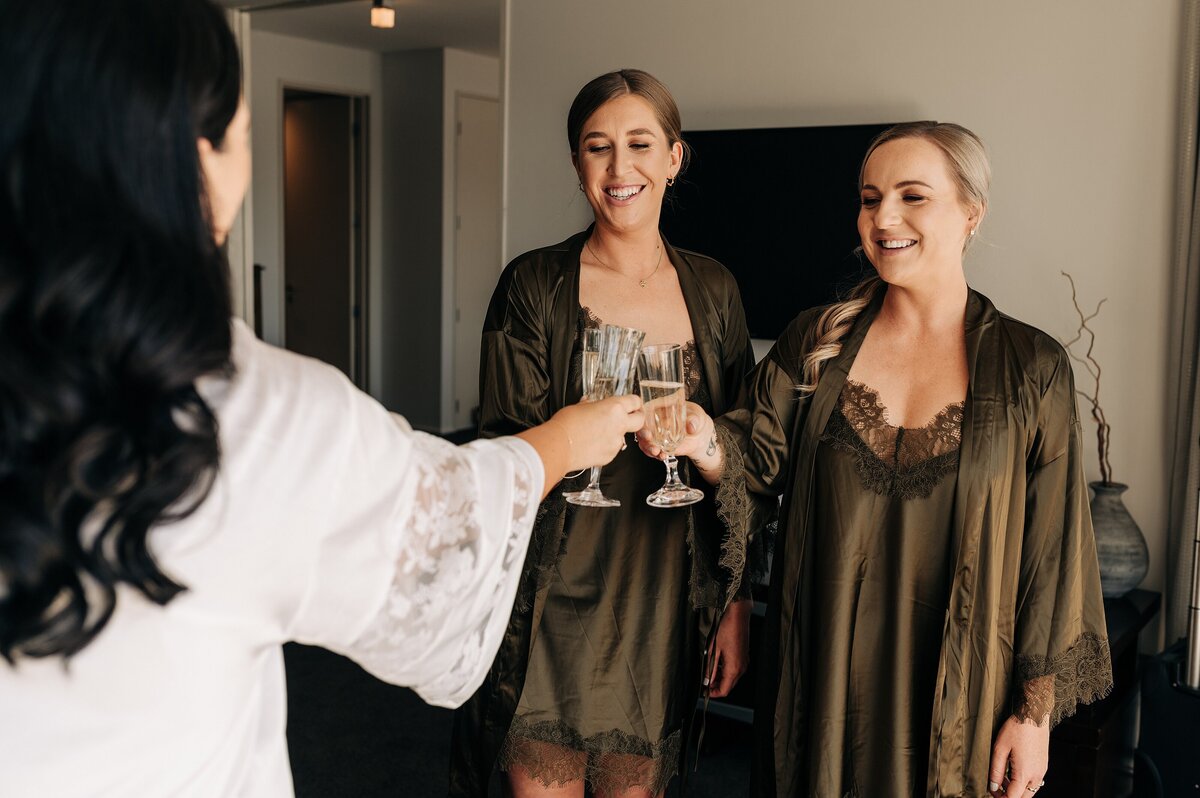 two bridesmaids in olive silk robes cheers with champagne with bride in white at clearwater resort christchurch before wedding