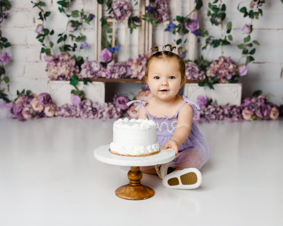 Baby in lavender dress holding a white cake during a cake smash session with purple floral backdrop. Photograph by Yucaipa cake smash photographer Kaitlyn Dawn Photography.
