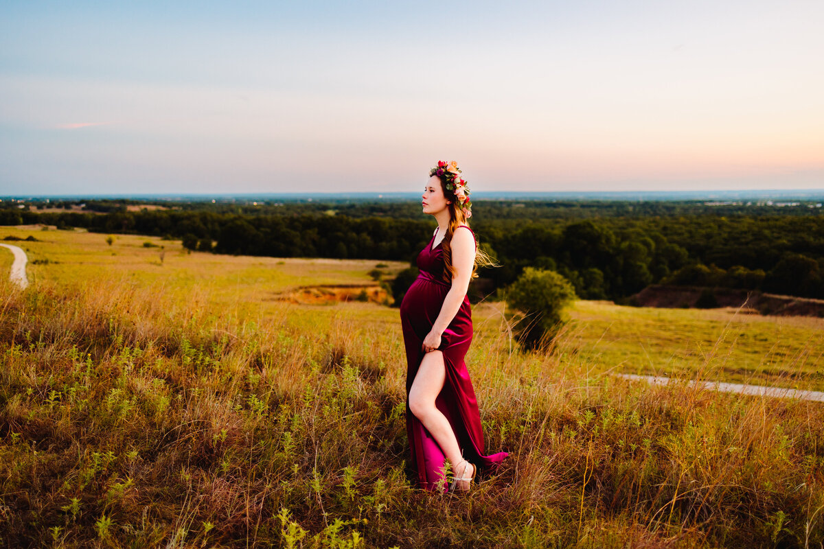 Affordable maternity photography in Albuquerque featuring a beautiful expectant mother wearing a long red dress and a flower crown. She poses gracefully on a green mountain, capturing the essence of natural beauty and anticipation.