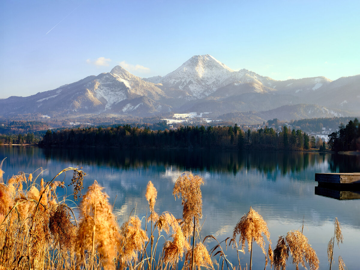 Stimmungsvolle Winteraufnahme vom Faaker See mit Blick über ruhiges Wasser, verschneite Berge und vereistes Schilf – Landschaftsfotografie aus Kärnten für Werbung und Editorials