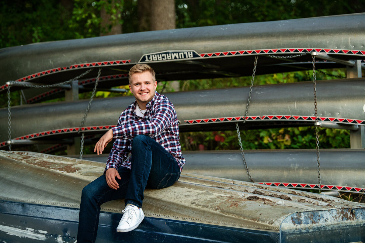 senior photo of guy sitting on canoe wearing plaid shirt and jeans