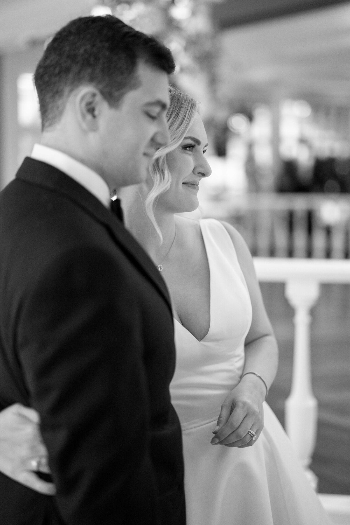 A black and white photo of a couple standing closely together. The woman wears a sleeveless dress and has a gentle smile. The man is in a suit. They appear to be attending a formal event indoors, with blurred decorations in the background.