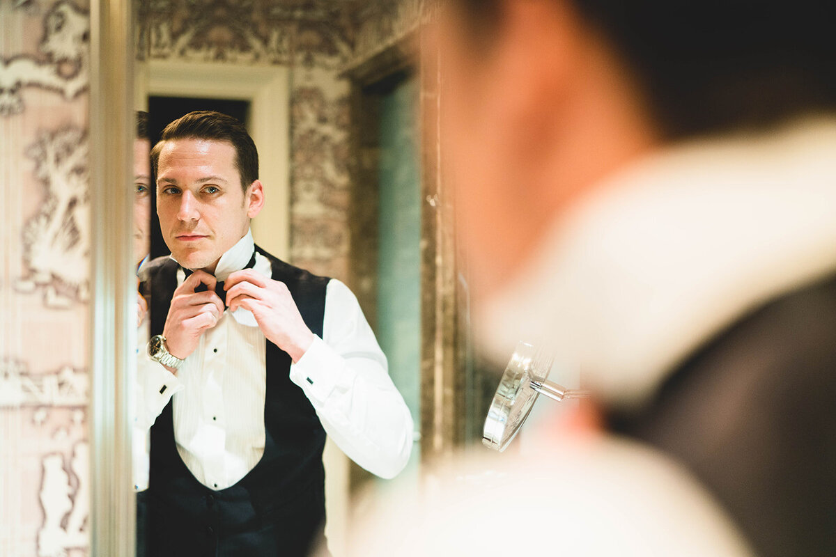 The groom is it doing his bow tie in front of the mirror