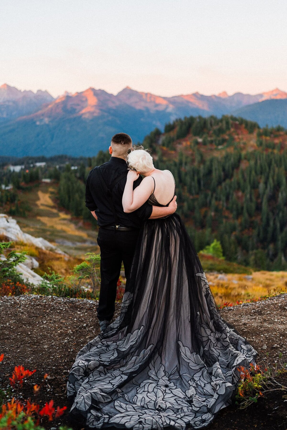 Bride with a long flowing black dress leaning into her groom watching the alpenglow of the north cascade mountains at the end of their elopement. 