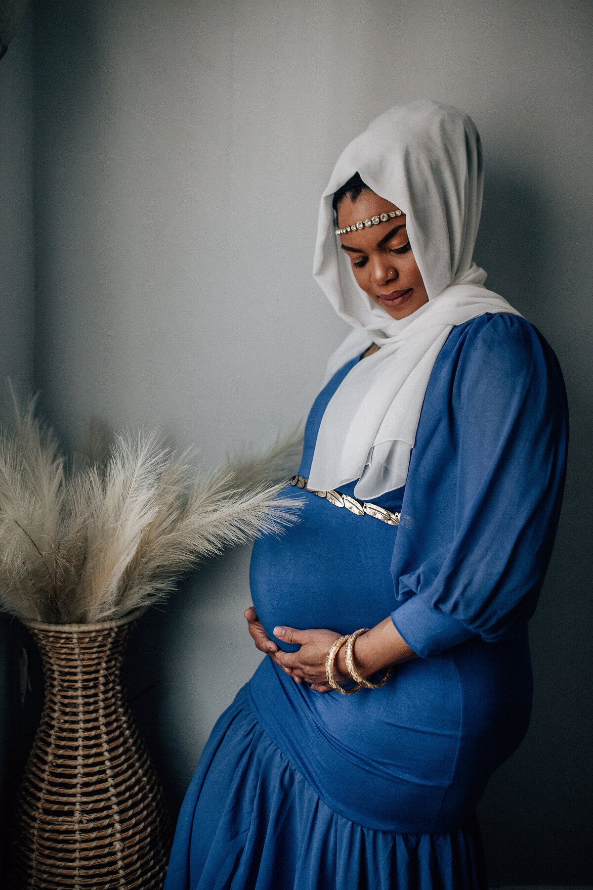 Pregnancy photoshoot with woman wearing blue dress, white scarf, and holding her belly.
