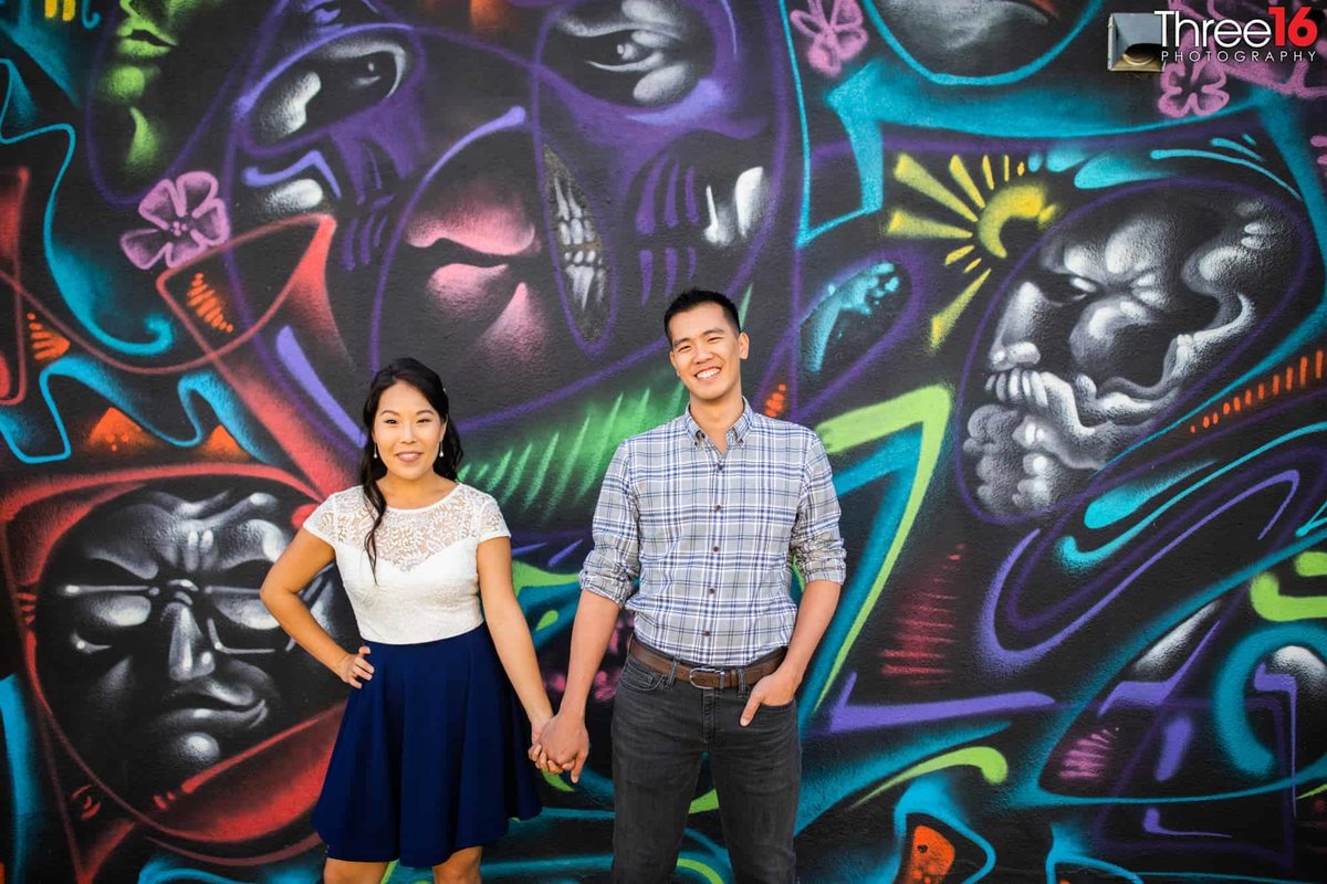 Engaged couple hold hands as they pose in front a large colorful mural in LA