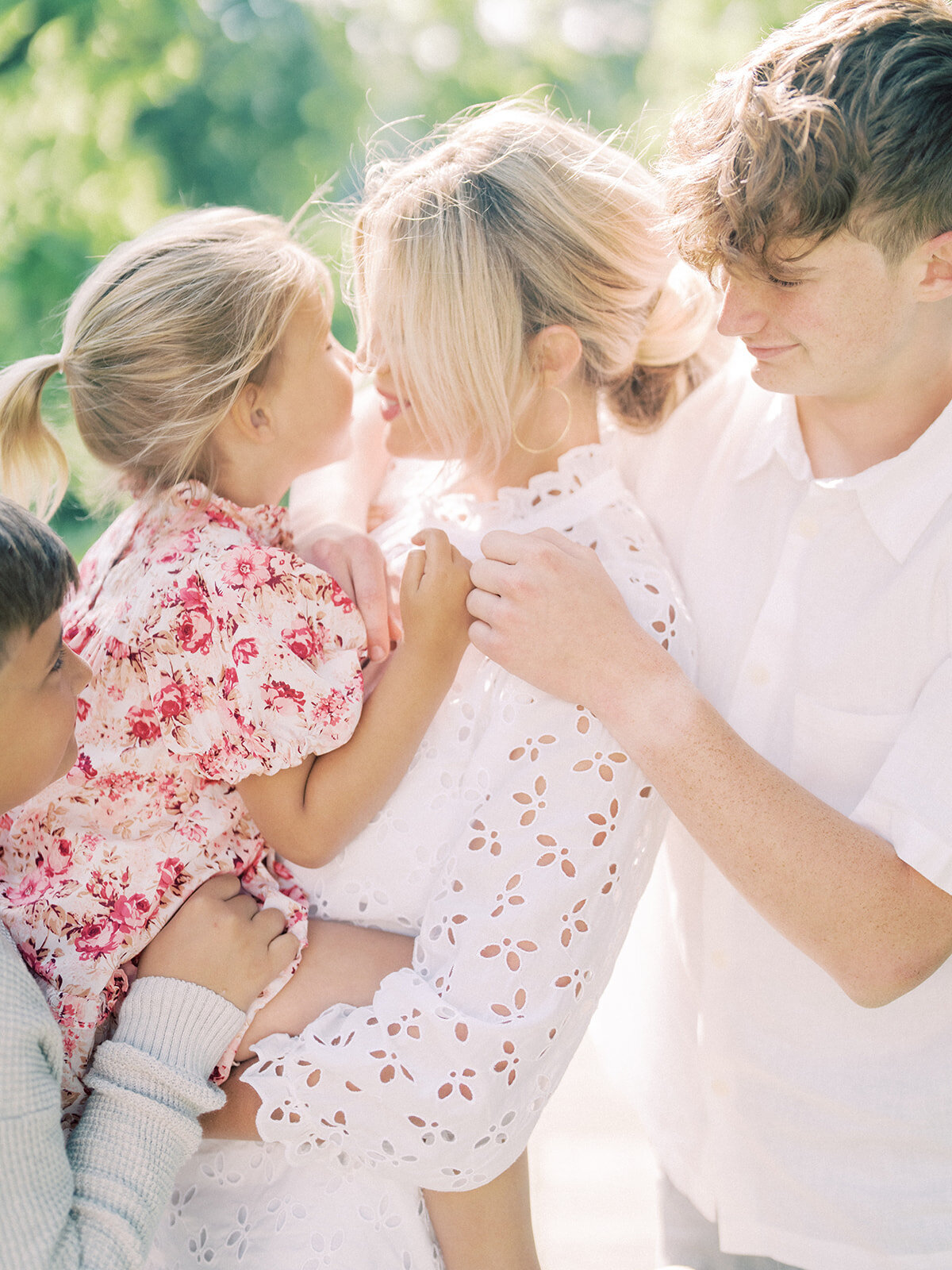 Little girl in floral pink dress is held by her mother and leans into her while older brothers stand on either side of mother.