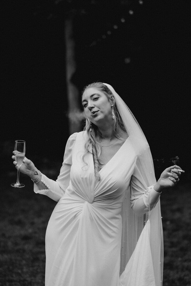 La mariée, une coupe de champagne dans une main et une cigarette dans l’autre, juste avant sa cérémonie laïque dans les bois de La Roche Couloir, en région parisienne.