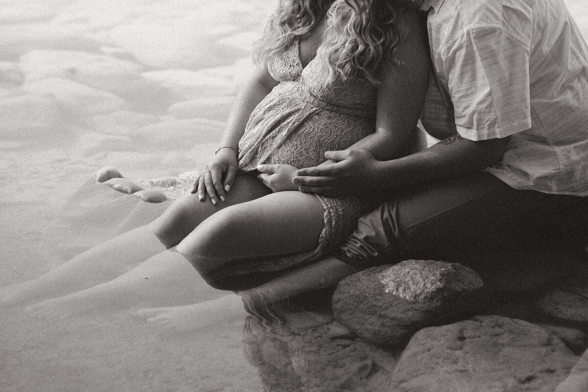A husband cradles his pregnant wife while they sit in the water. This black and white portrait taken at Johnson Lake by Brittany Danielle