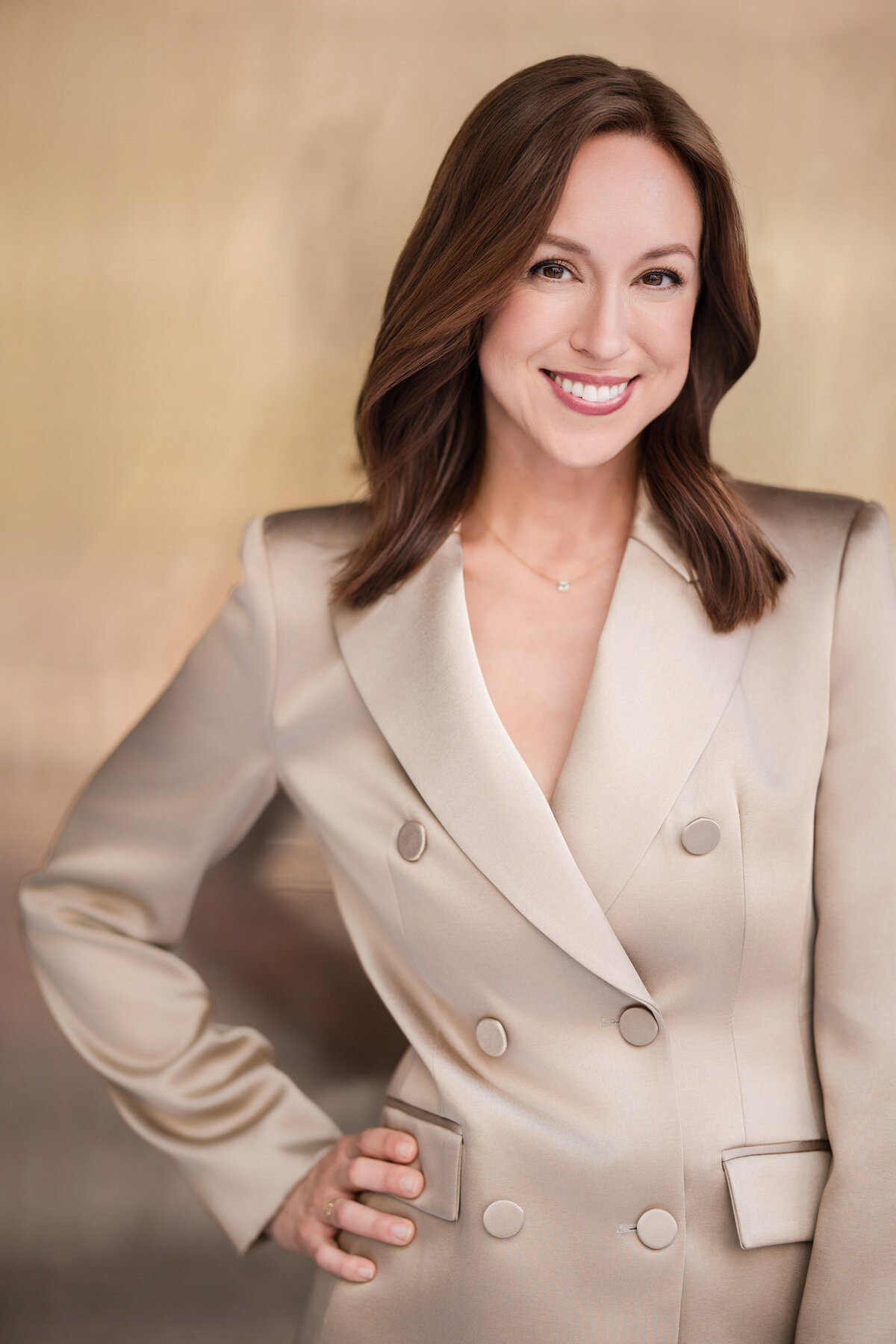 Person standing confidently in front of a warm-toned background, wearing a tailored, double-breasted beige blazer. They have shoulder-length brown hair styled in soft waves, a natural makeup look, and are smiling directly at the camera with one hand on their hip.