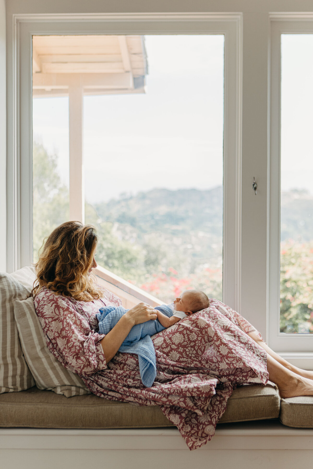 Mom sits by the window with view of Los Angeles while crading newborn baby in her lap.