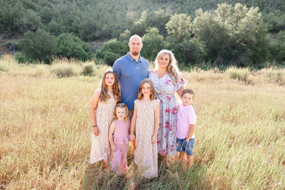 A family of six stands in a sunlit field in Midway, Utah, dressed in coordinating pastel outfits. The parents and their four children smile warmly as they pose against a scenic backdrop of rolling hills and greenery.