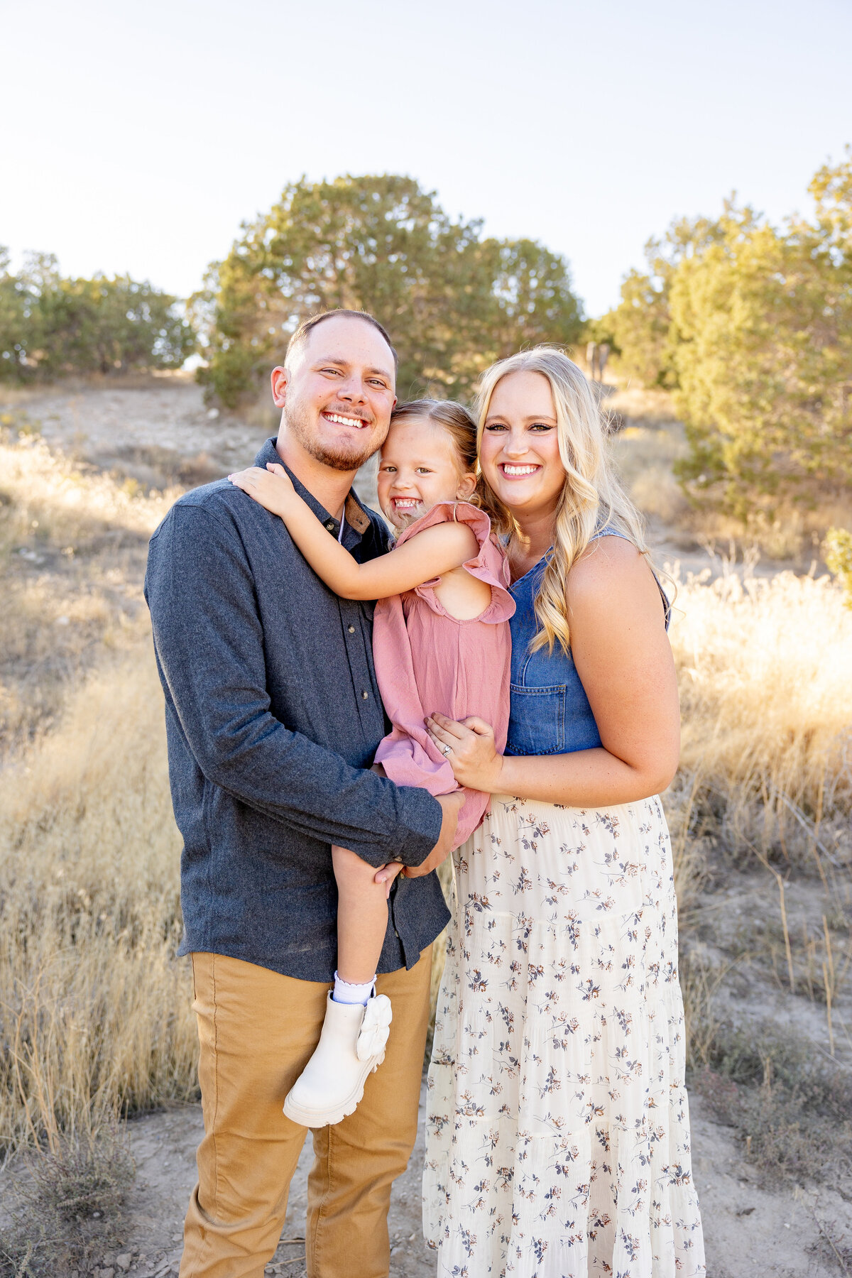 A mother and father hold their daughter close while standing in the golden fields of Eagle Mountain, Utah. They are all smiling, radiating love and joy in the soft sunlight.
