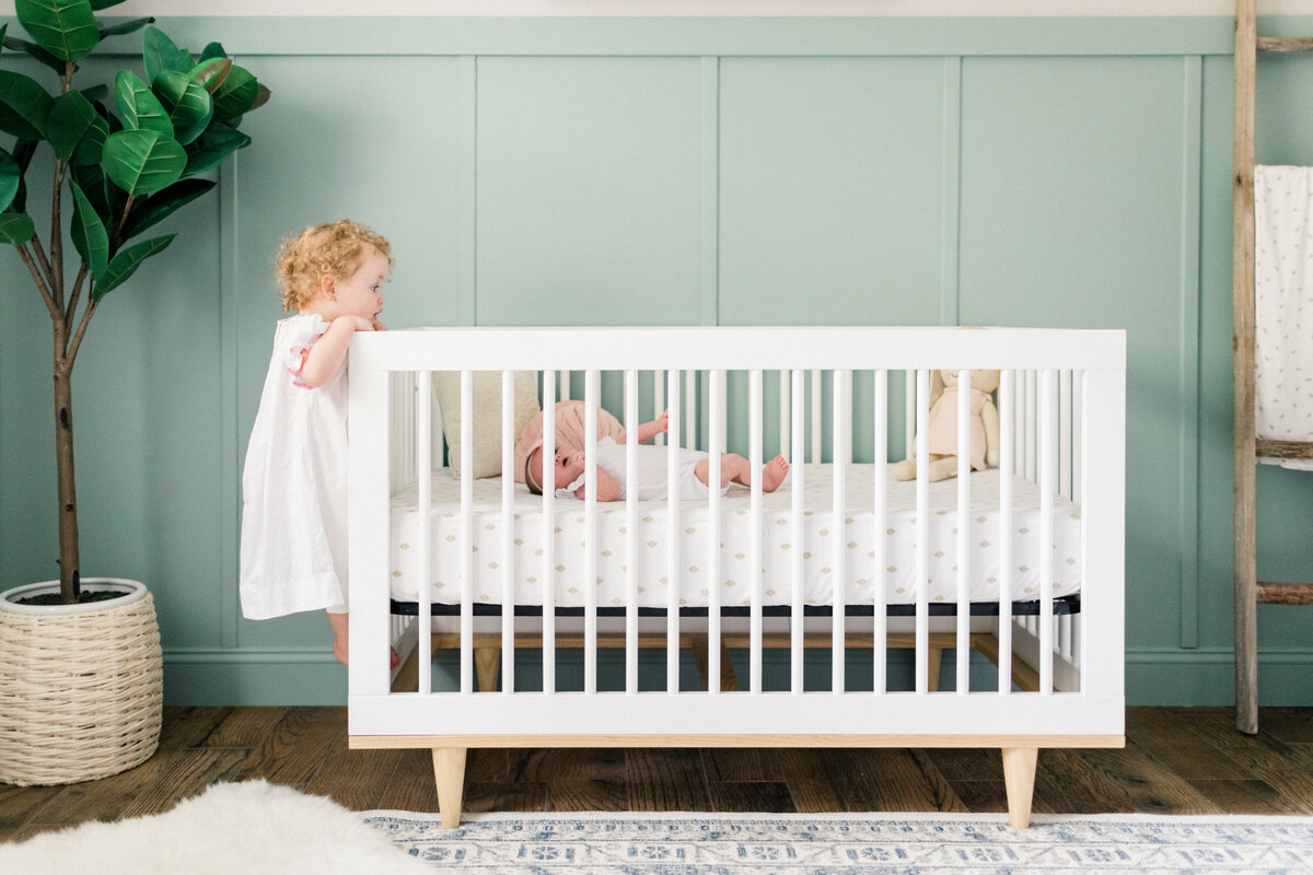 toddler peeking at new baby over crib by Orlando in-home newborn photographer