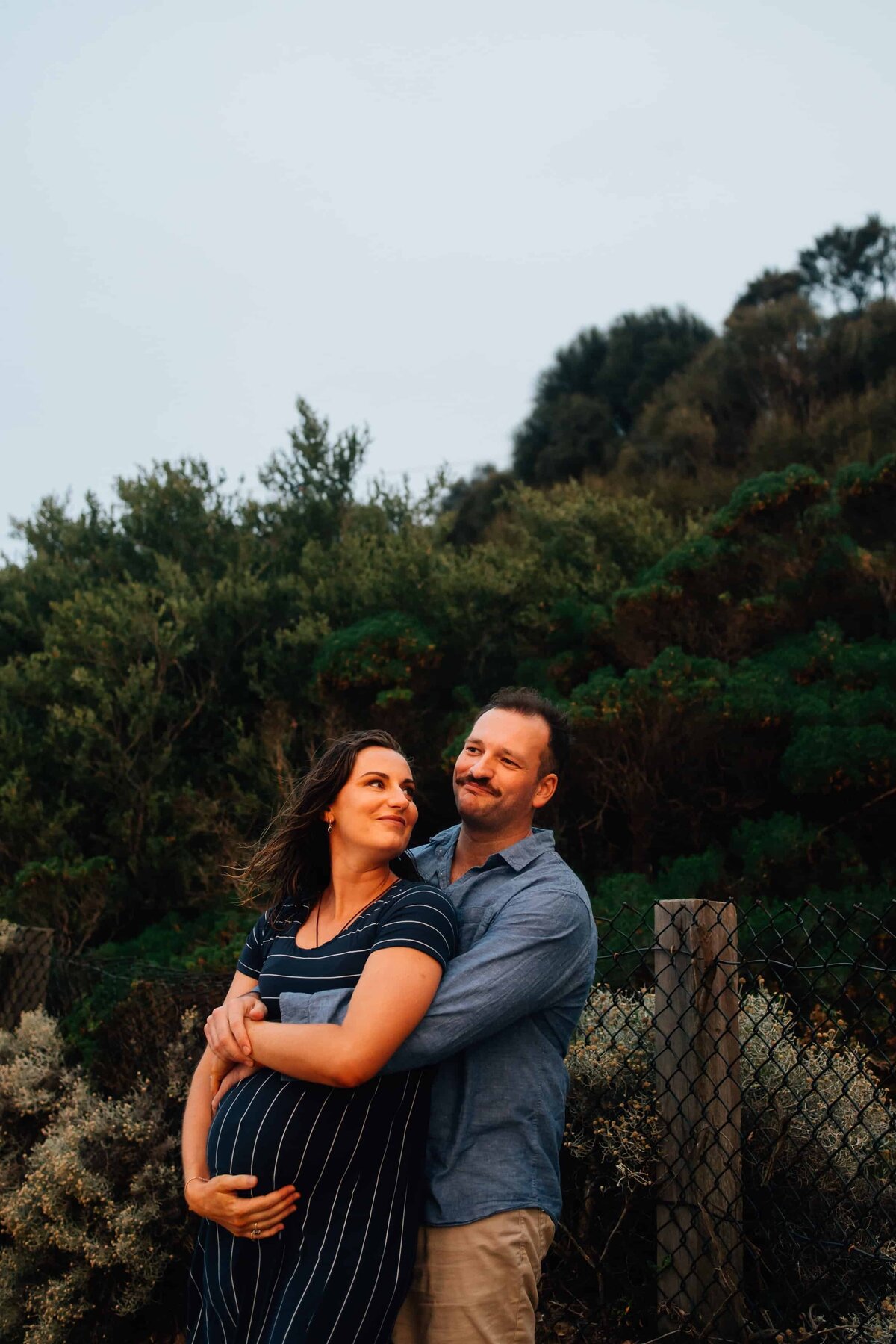 Man and woman standing, his arms around her, on beach maternity photoshoot Melbourne