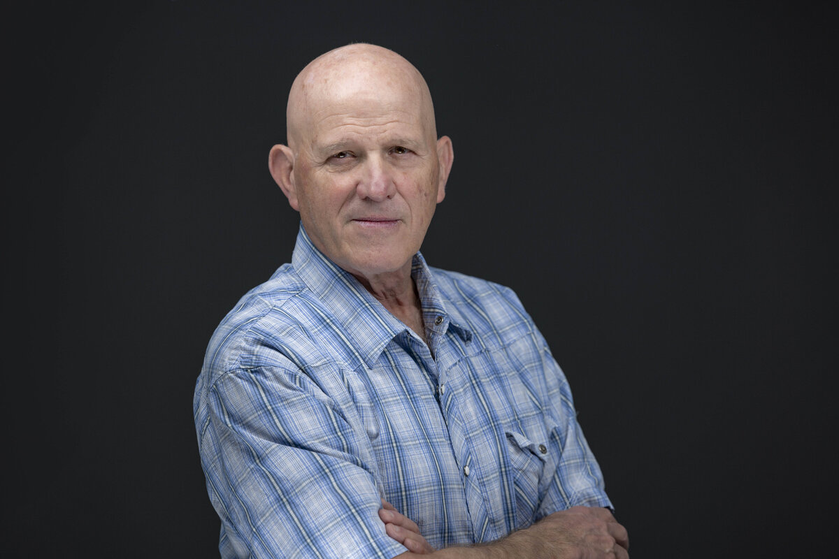 Business headshot in CT of a bald man wearing a light blue checkered shirt, arms crossed, photographed against a dark backdrop by Rob Faber Photography, expert in headshots in CT and MA.