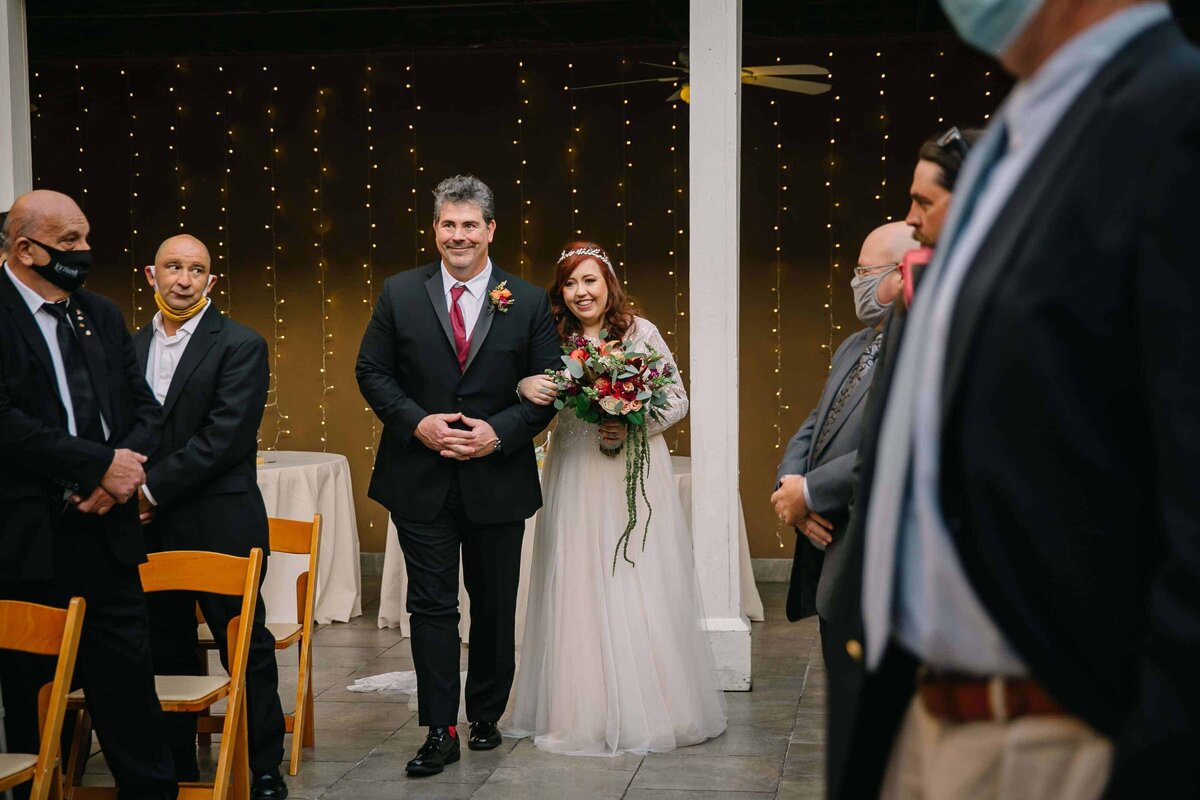 A bride in a white dress holding a bouquet walks down the aisle with a man in a suit, surrounded by seated and standing guests, some of whom are wearing masks. String lights hang in the background.