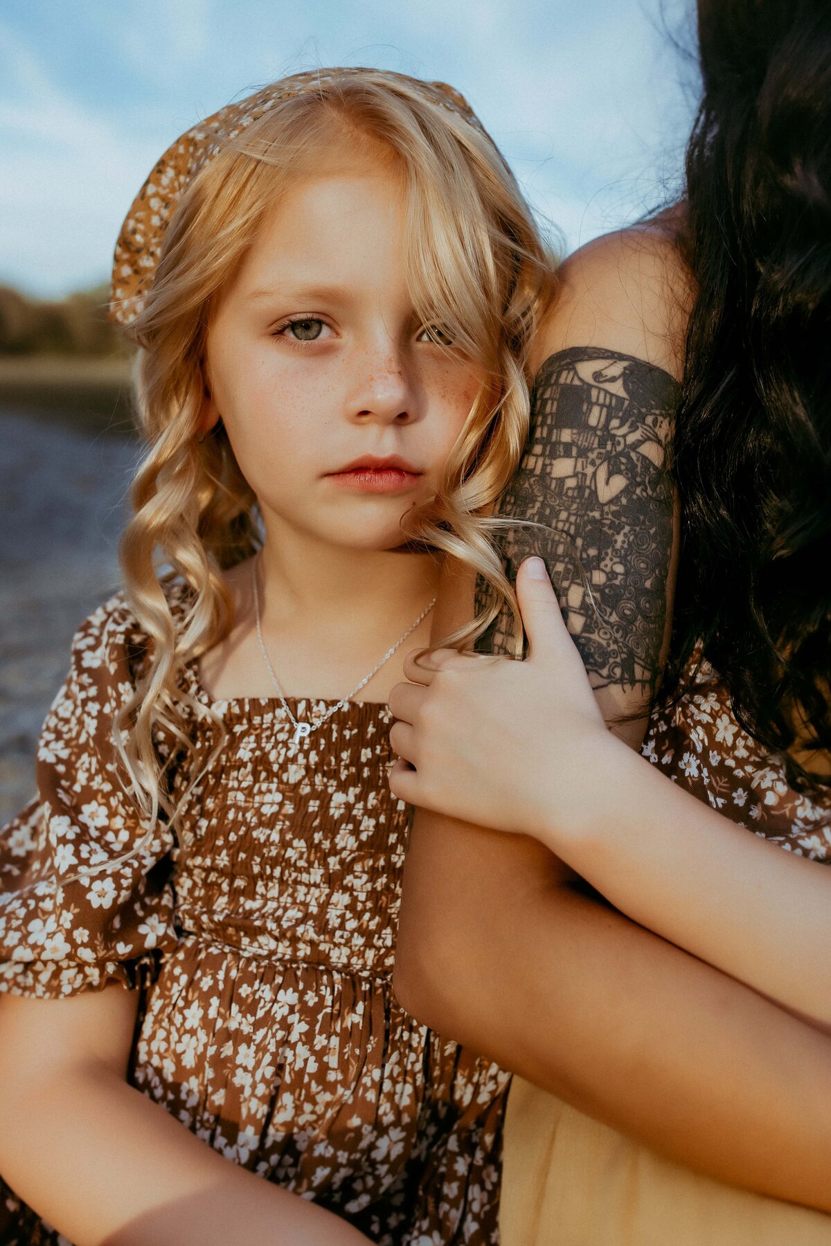 A young girl with blonde hair in a floral dress holds the arm of a tattooed individual, both captured beautifully by a photographer in Warner Robins, GA, as they sit outdoors.