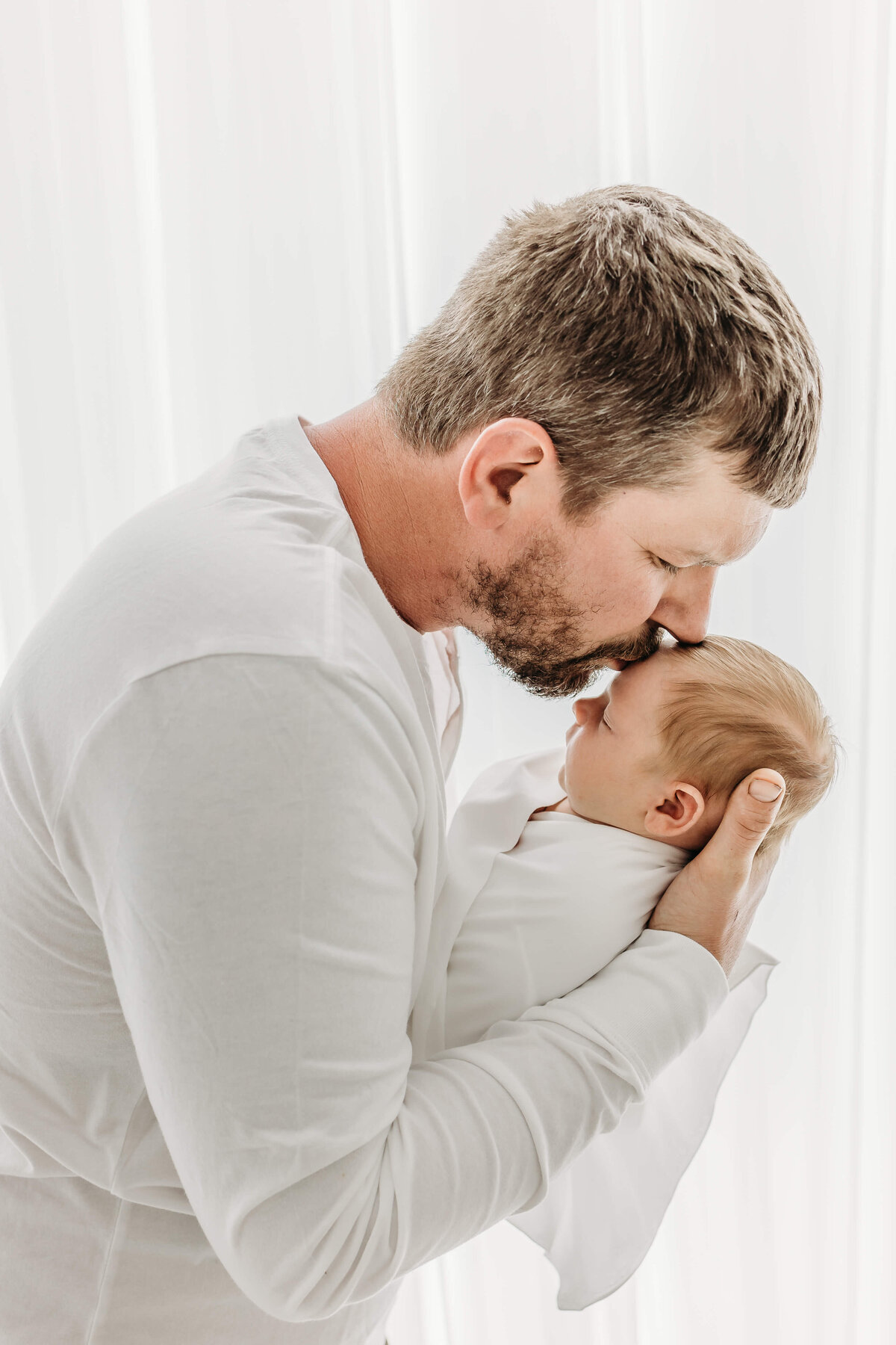 Dad in white shirt holding wrapped newborn and kissing head