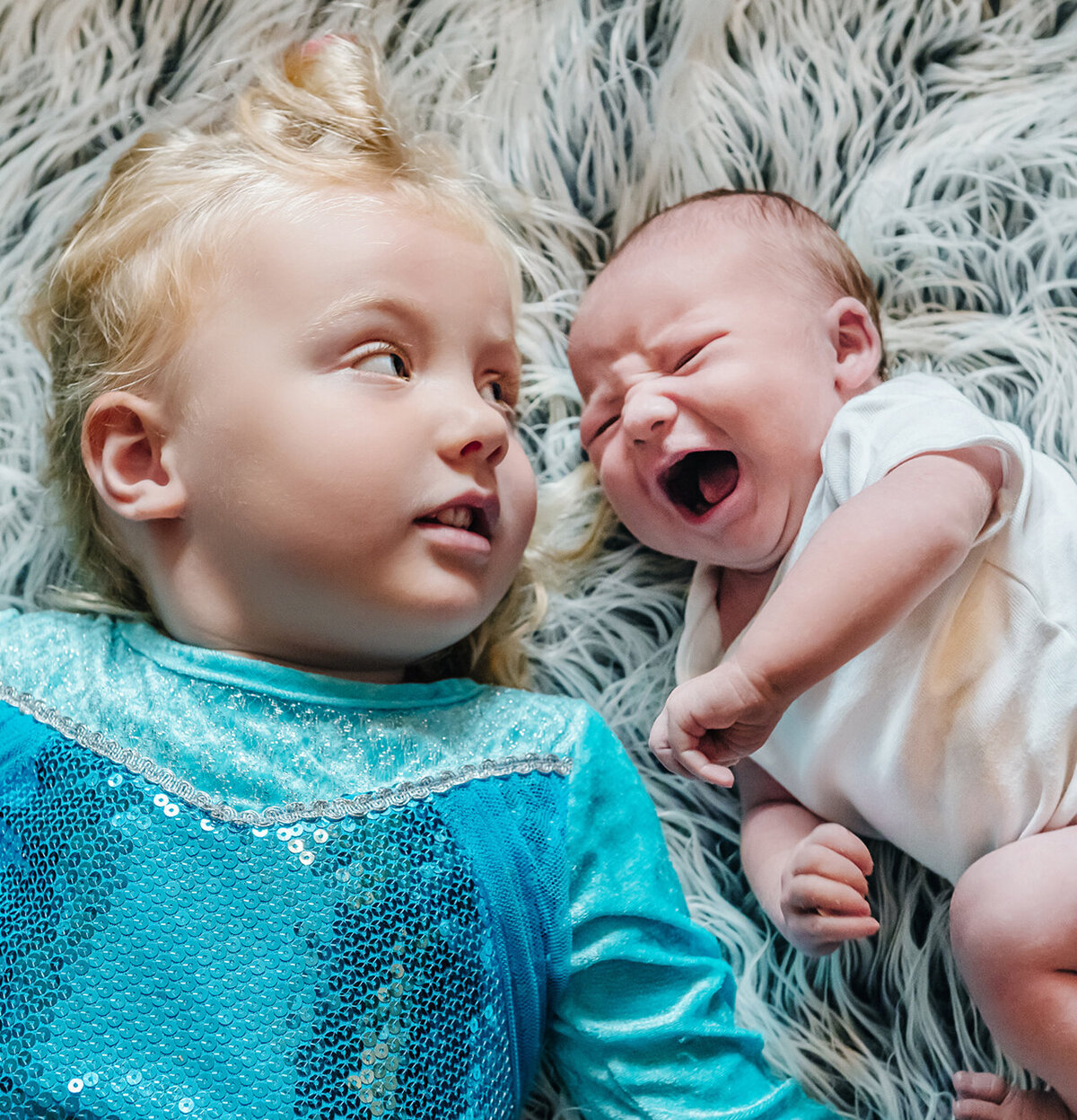 Big sister while wearing a Frozen dress looks at her newborn sister who is crying while they both lie on a fuzzy carpet.