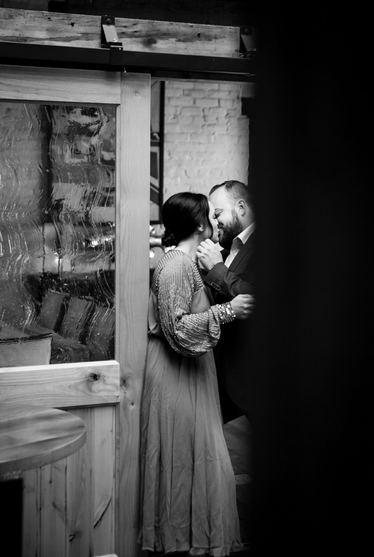 The camera peeks around a wall at a man leaning in for a kiss with his fiancée.