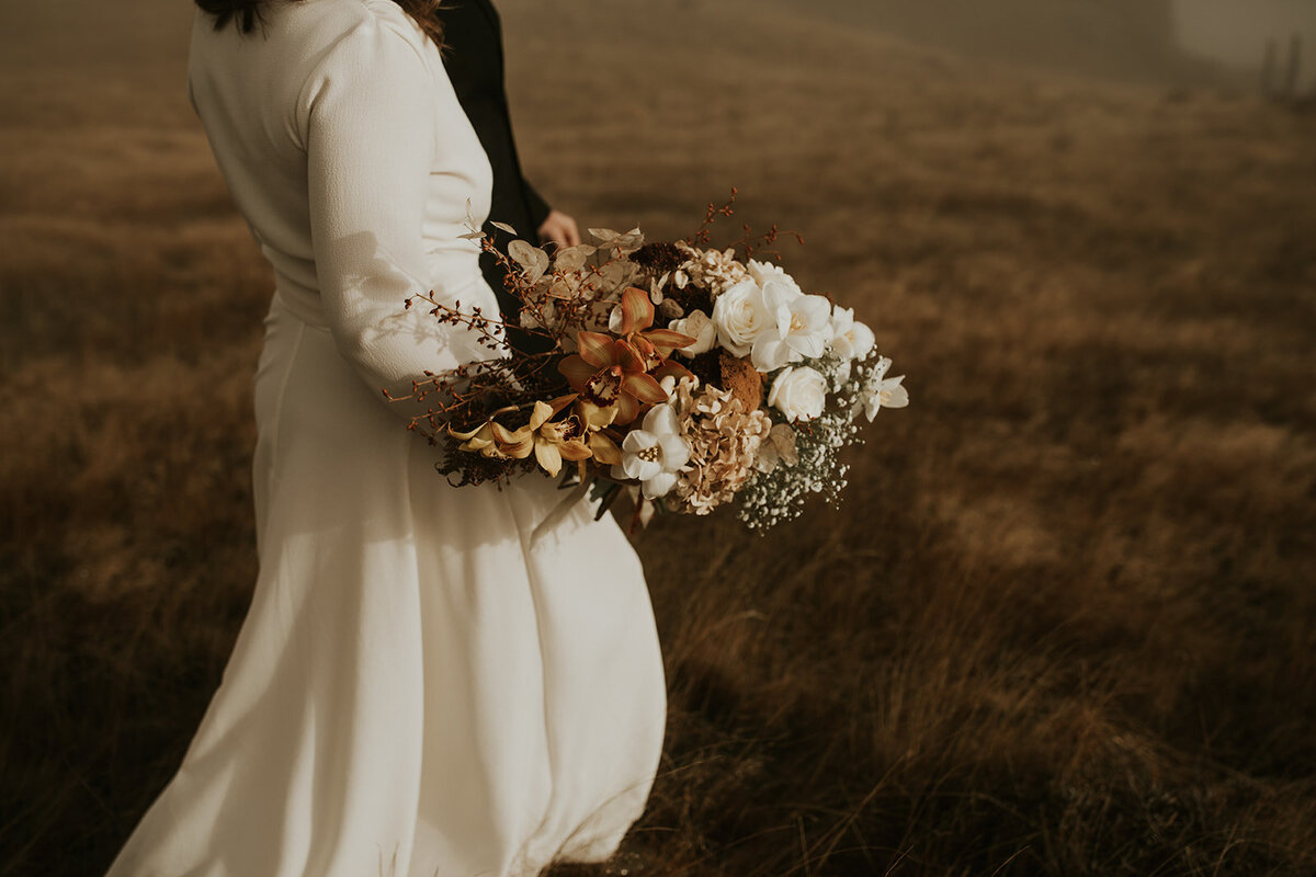 The Vase Floral Co - Bridal bouquet held as bride walks through New Zealand landscape