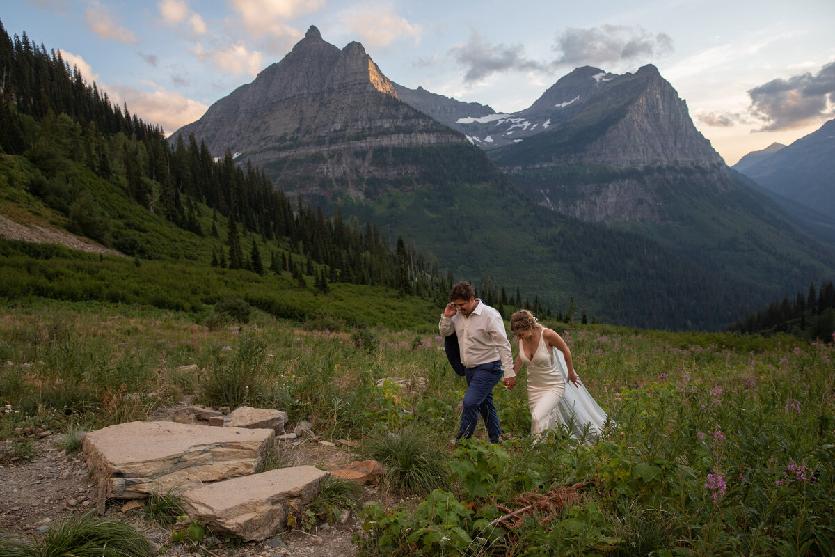 A bride and groom walk hand in hand through a meadow with wildflowers as the sunsets in the mountains behind them.