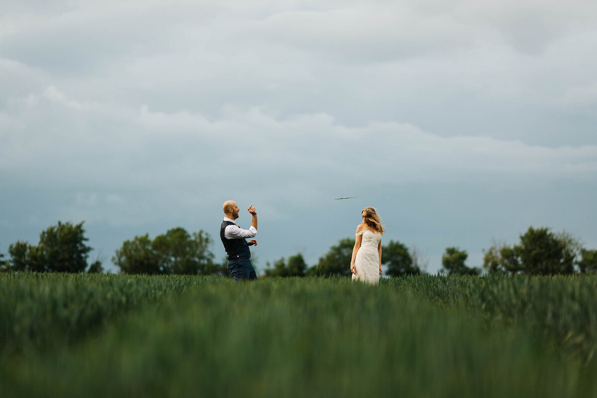 Bride and groom stood in a corn field with the groom throwing a piece of grass like a paper airplane