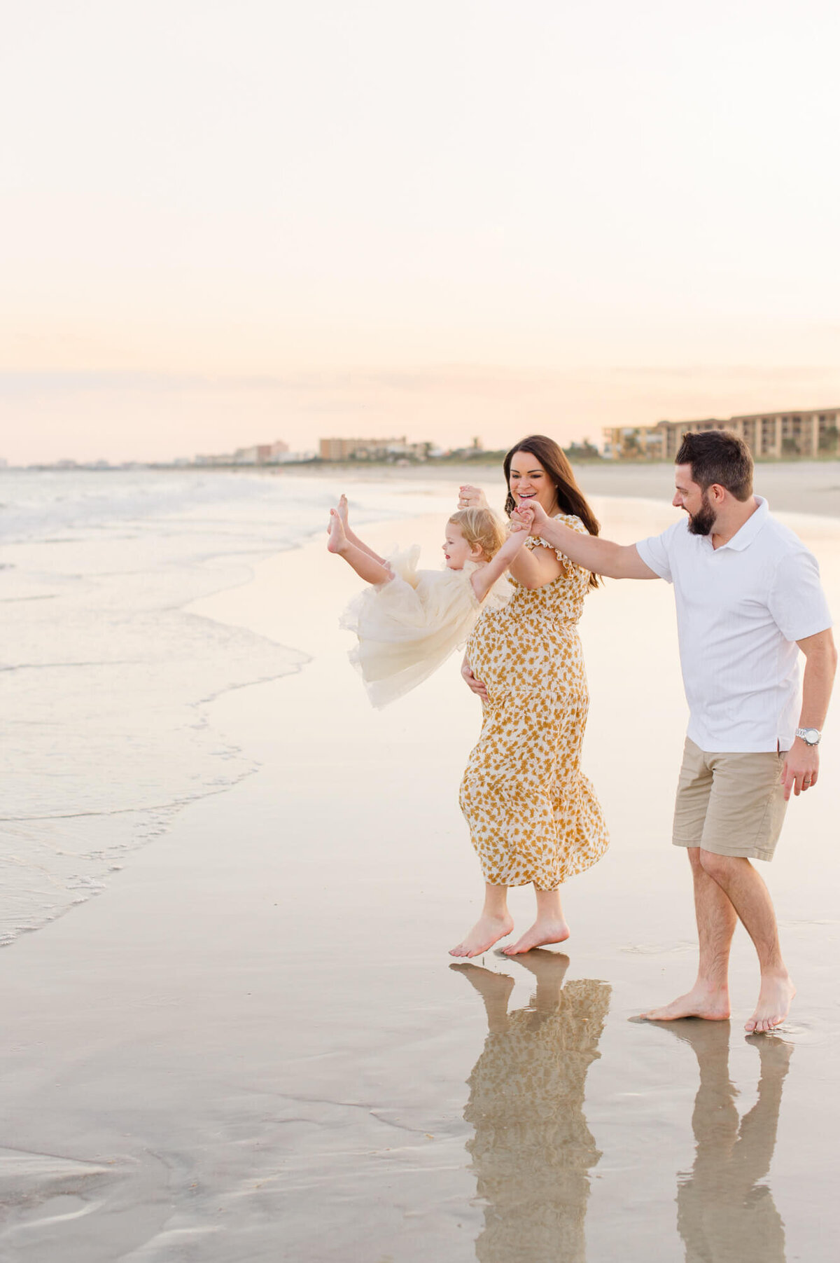 Expectant parents walk along beach at the shoreline swinging their toddler daughter in the air while she laughs at sunset