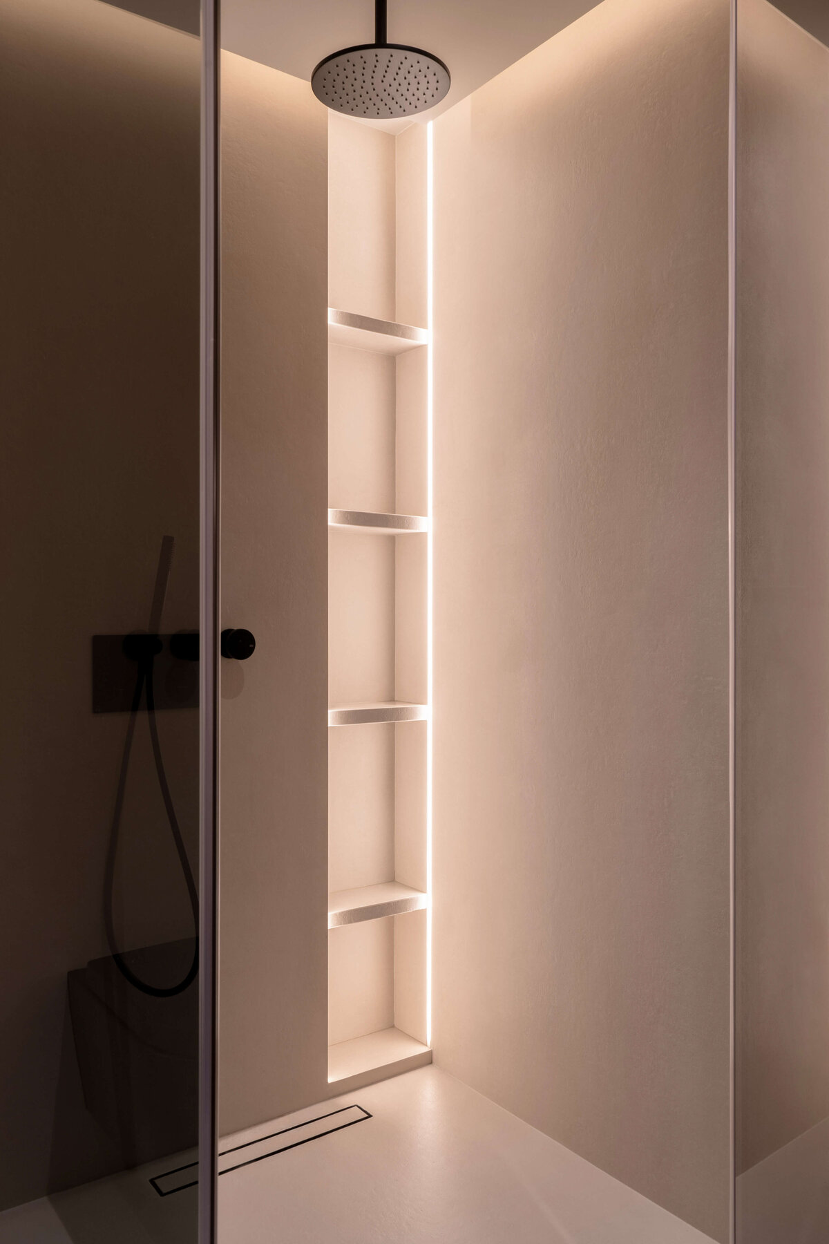 Large, minimal walk in shower. Smokey black glass and black shower hardware provide a dramatic look against the otherwise light and airy space. Built-in shelving within the shower emits a soft light.