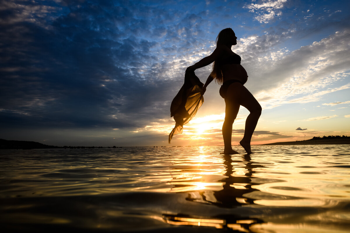 A dramatic maternity silhouette photo of a pregnant woman standing barefoot in shallow water, holding a flowing scarf as the sun sets brightly behind her, casting a golden reflection on the water and sky