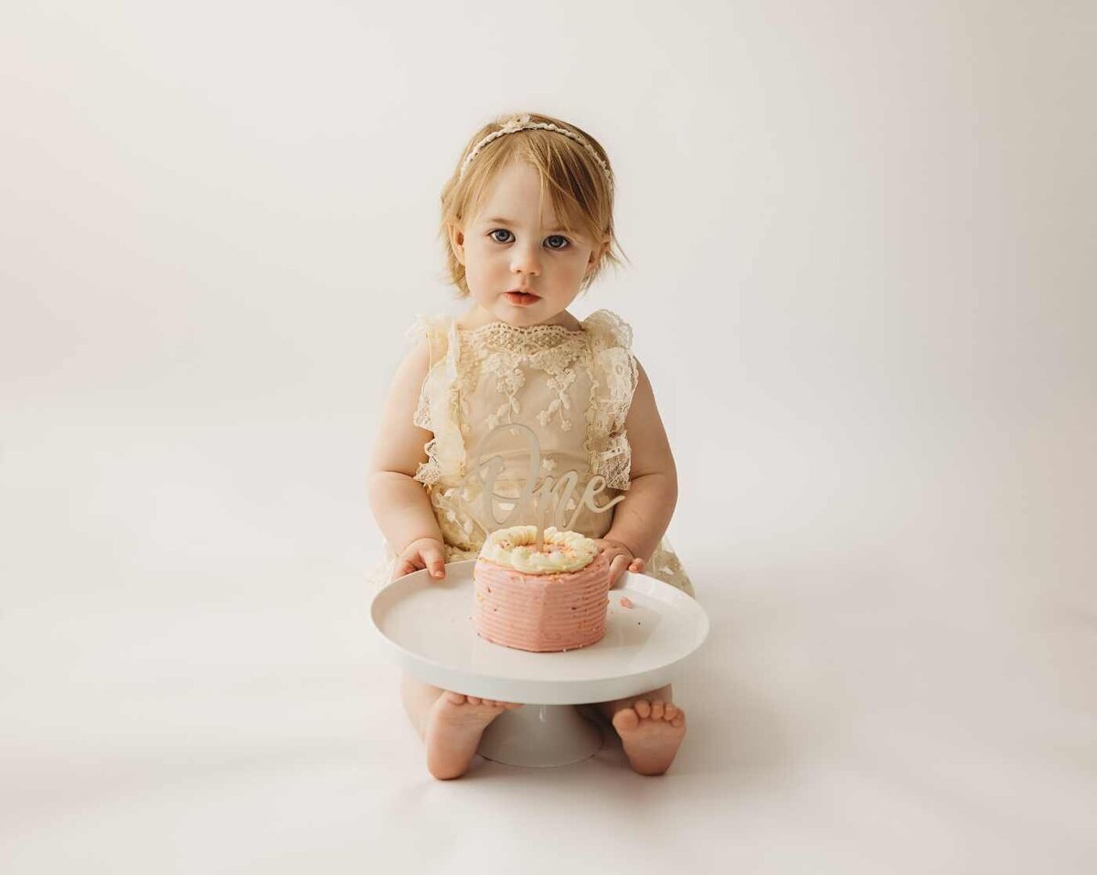 Ruua Hamdan Photography (UK): A baby wearing a "one" dress sits holding a small pink and white cake on a white plate.