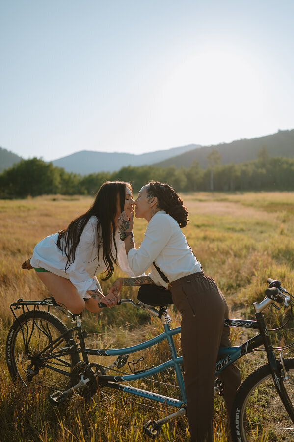 A woman kisses her fiancé who is balancing on the backside of a bicycle. They're in a grassy field somewhere in Idaho Falls, ID.