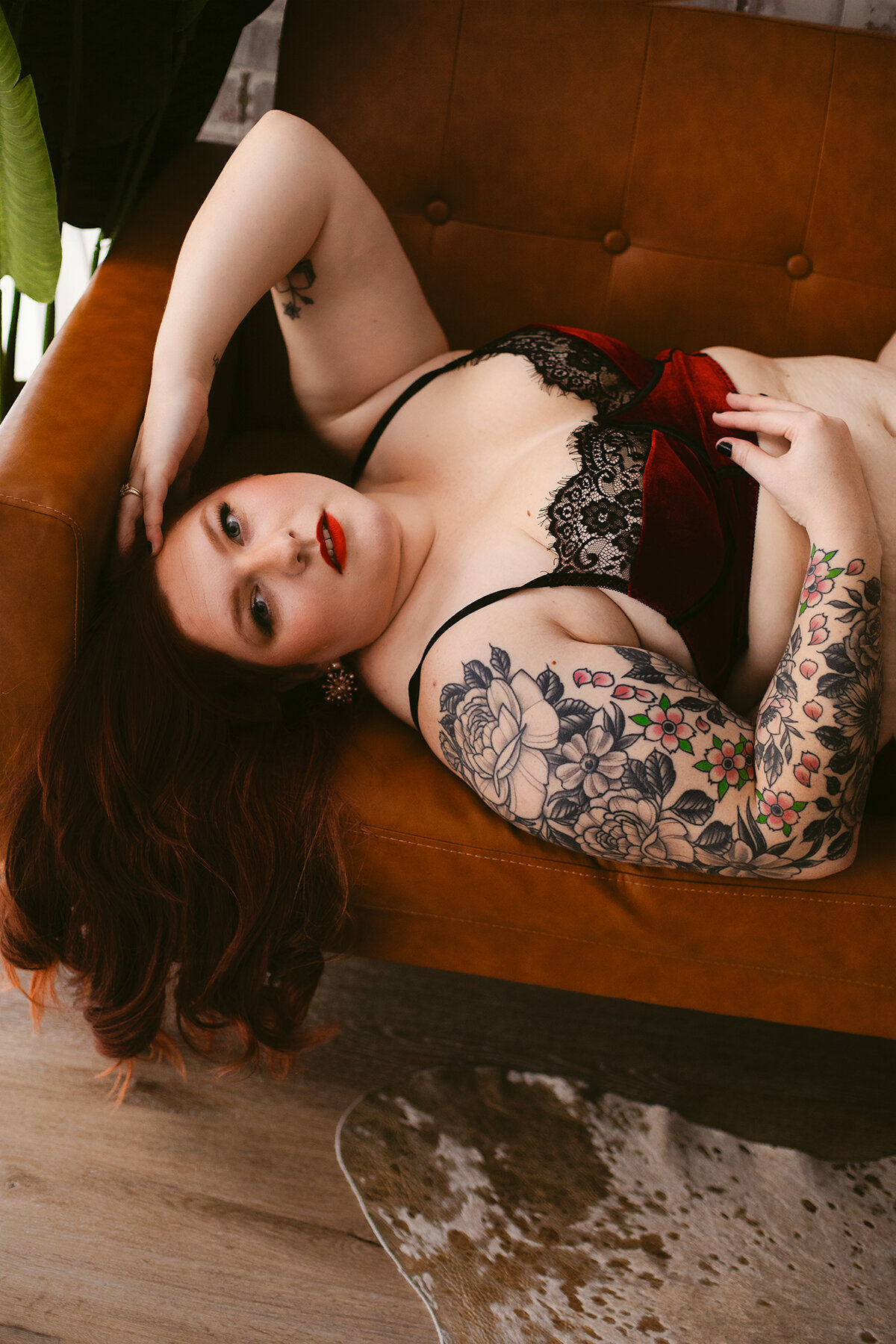 A woman in red linger lays on a tan couch with her hand in her hair for a boudoir photoshoot.