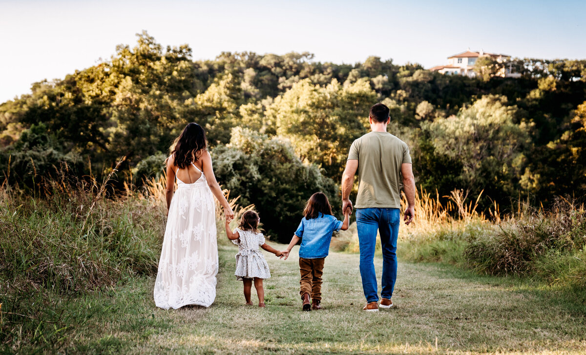 Family Photography, a young family walks hand-in-hand on a grassy trail toward the forest