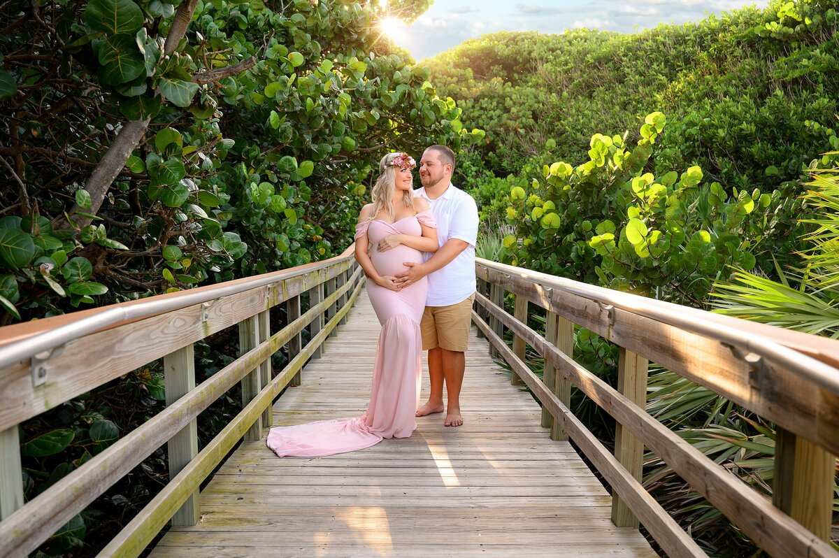 Pregnant couple expecting their first baby posed on a boardwalk at the beach with a greenery background in the sunset