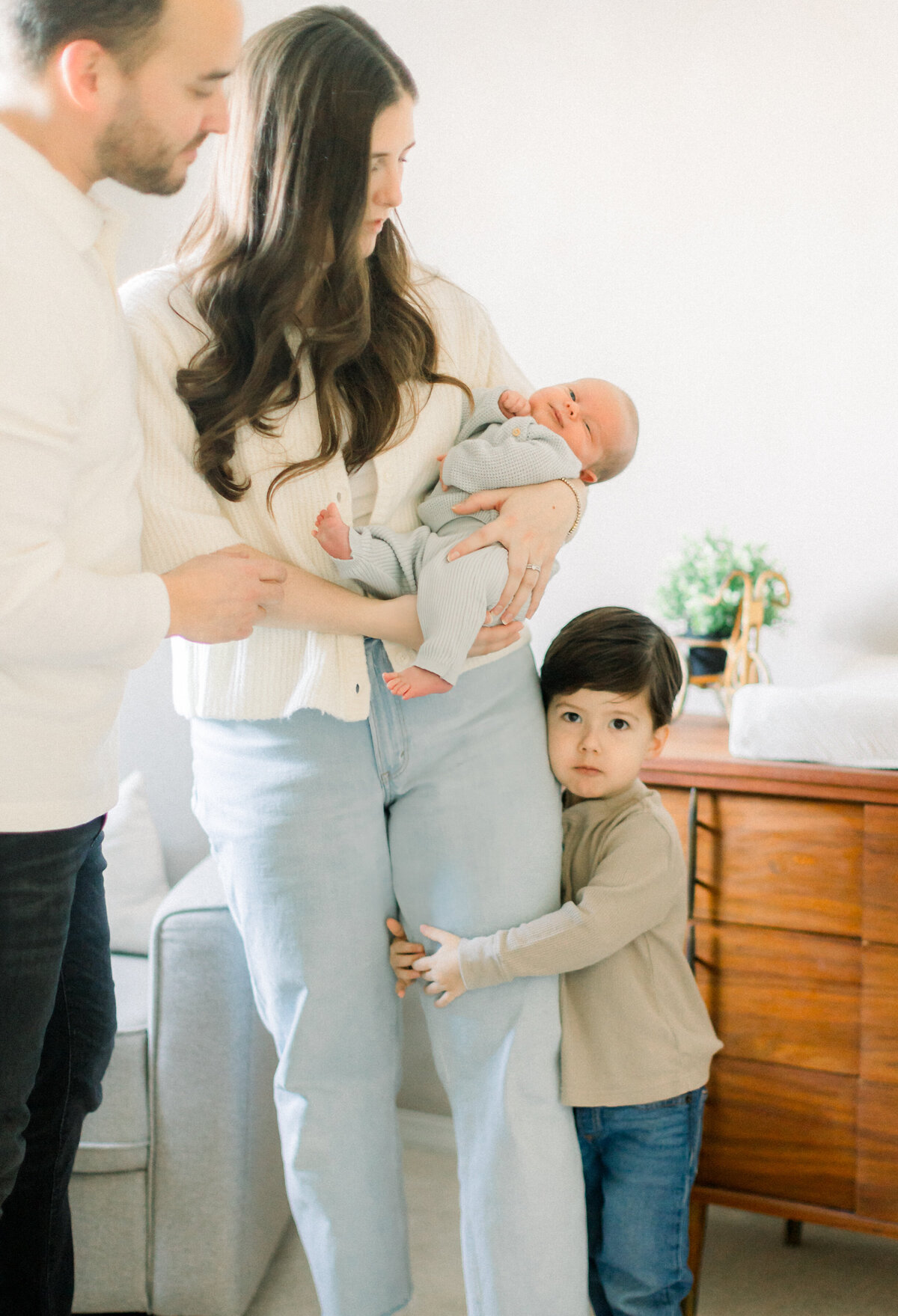 Mom holds newborn baby as young boy clings to her leg.