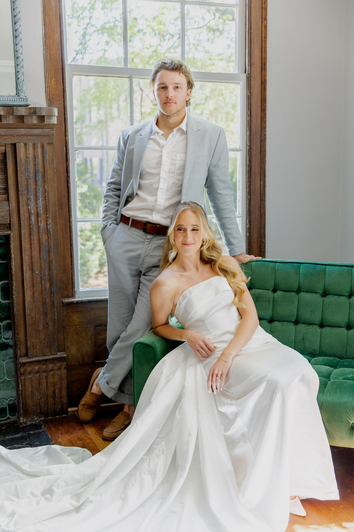 A bride sits on a forest green couch while her groom stands behind with his hand on the couch. The photo is very posed and polished.