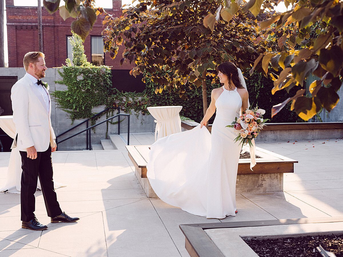 The groom, wearing a tuxedo with a white jacket, watches as the bride twirls in her silk sheath halter top wedding dress in the courtyard of Clementine Hall at their summer wedding. The bride is holding her tropical bouquet of flowers and palm fronds accented with long raw silk ribbons