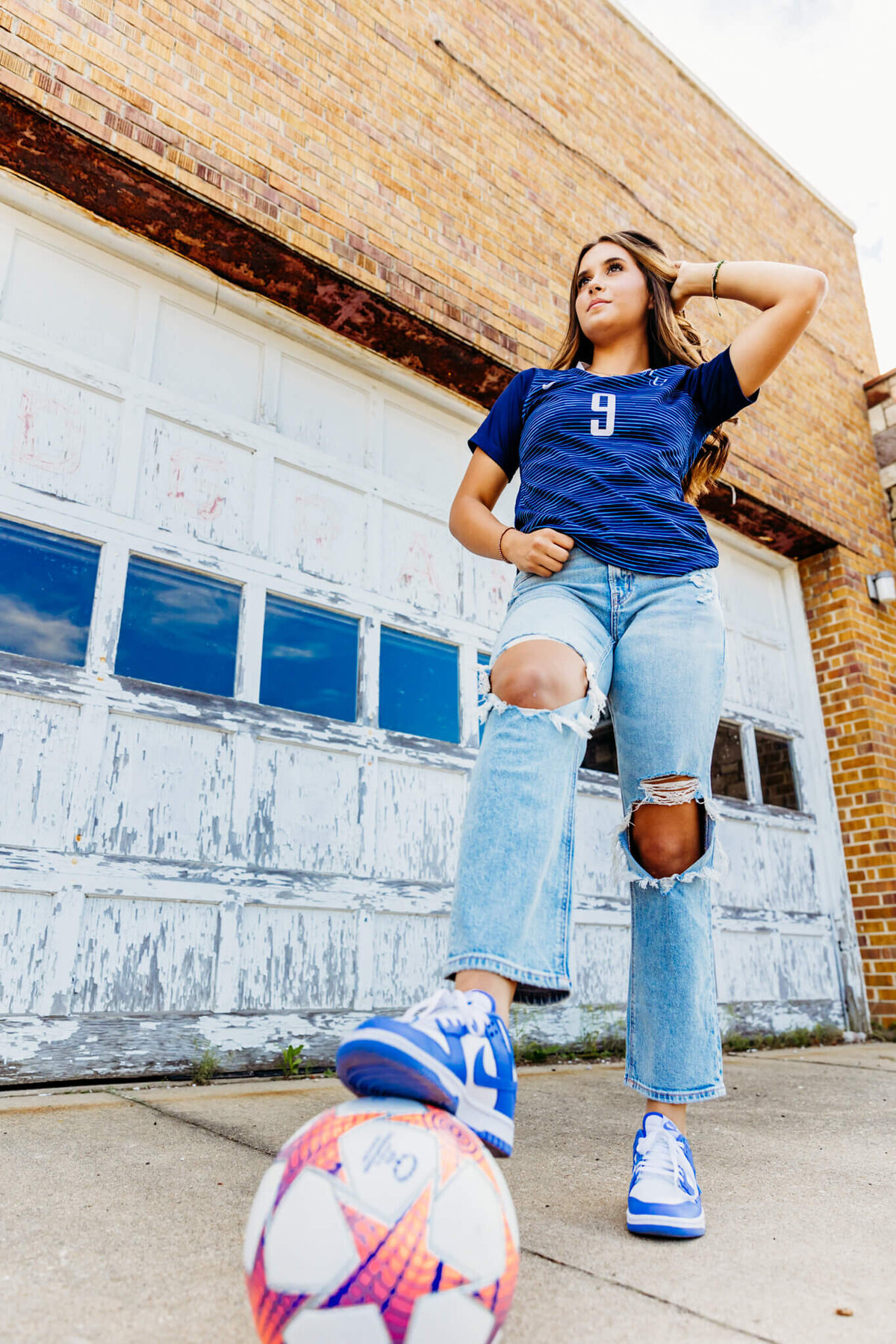 beautiful teenage high school girl in ripped jeans and blue soccer jersey resting her foot on a soccer ball as she stands in front of a rustic garage door near Oshkosh 