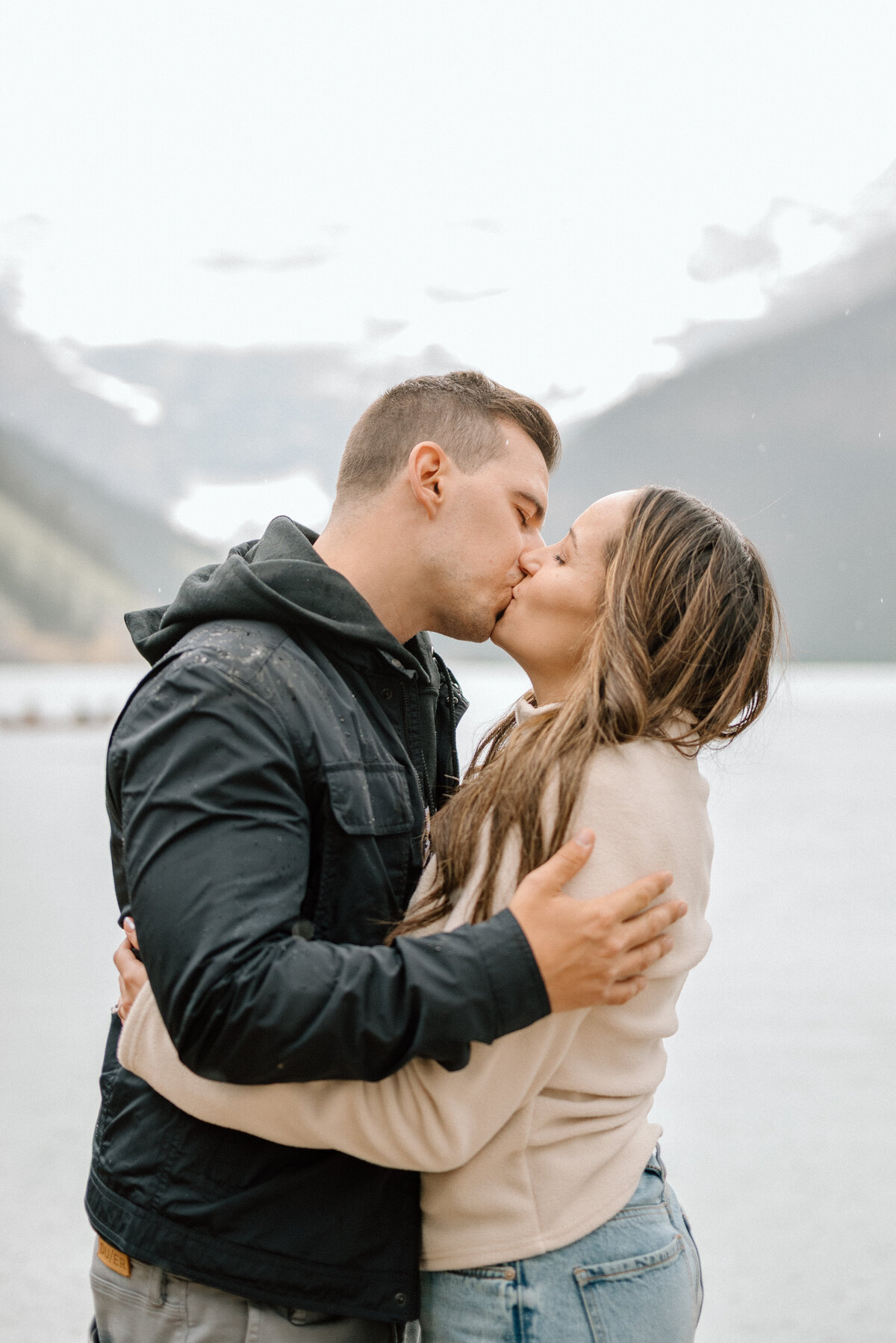 Close-up kiss by Lake Louise with misty peaks and soft light.