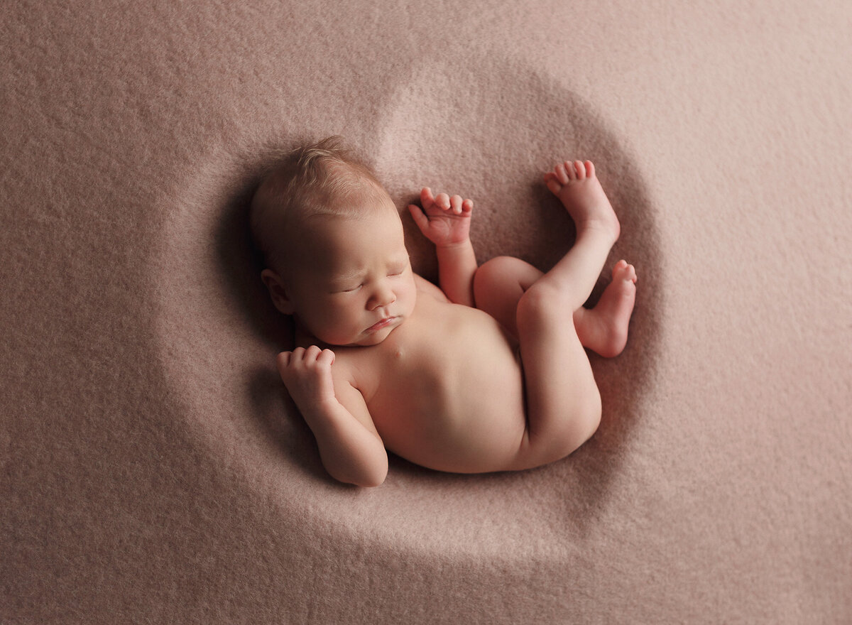 newborn baby girl posed on her back all curled up inside a wooden heart bowl that has been silhouetted by a soft pink textured backdrop