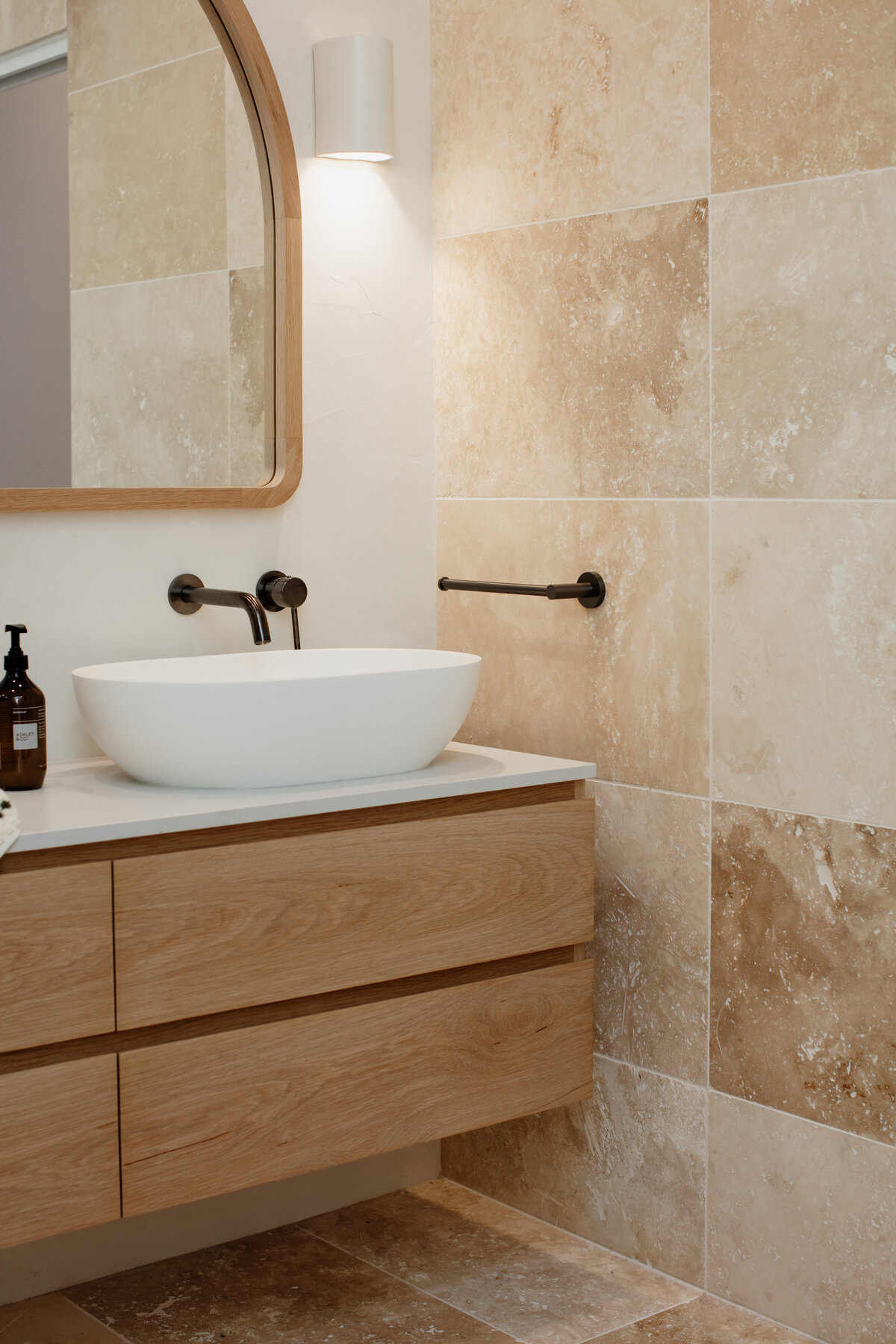 Modern bathroom crafted by a Newcastle Interior Designer, featuring a wooden vanity, white vessel sink, and beige stone tiles. The elegant wall-mounted faucet complements the illuminated mirror.